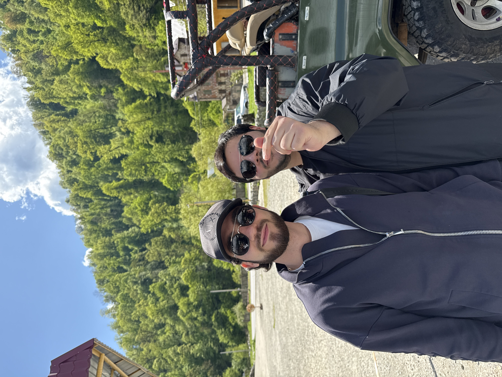 Two men are posing for a photograph in Teghut, Armenia. The background features lush green trees and a blue sky. The man in the foreground is wearing a navy jacket, a white t-shirt, and a baseball cap, along with sunglasses. He has a beard. The man behind him is wearing a black jacket and sunglasses and is making a finger heart gesture. They appear to be standing next to a dark green jeep with a visible tire. A building with a red roof is partially visible in the lower left corner. The ground appears to be paved.