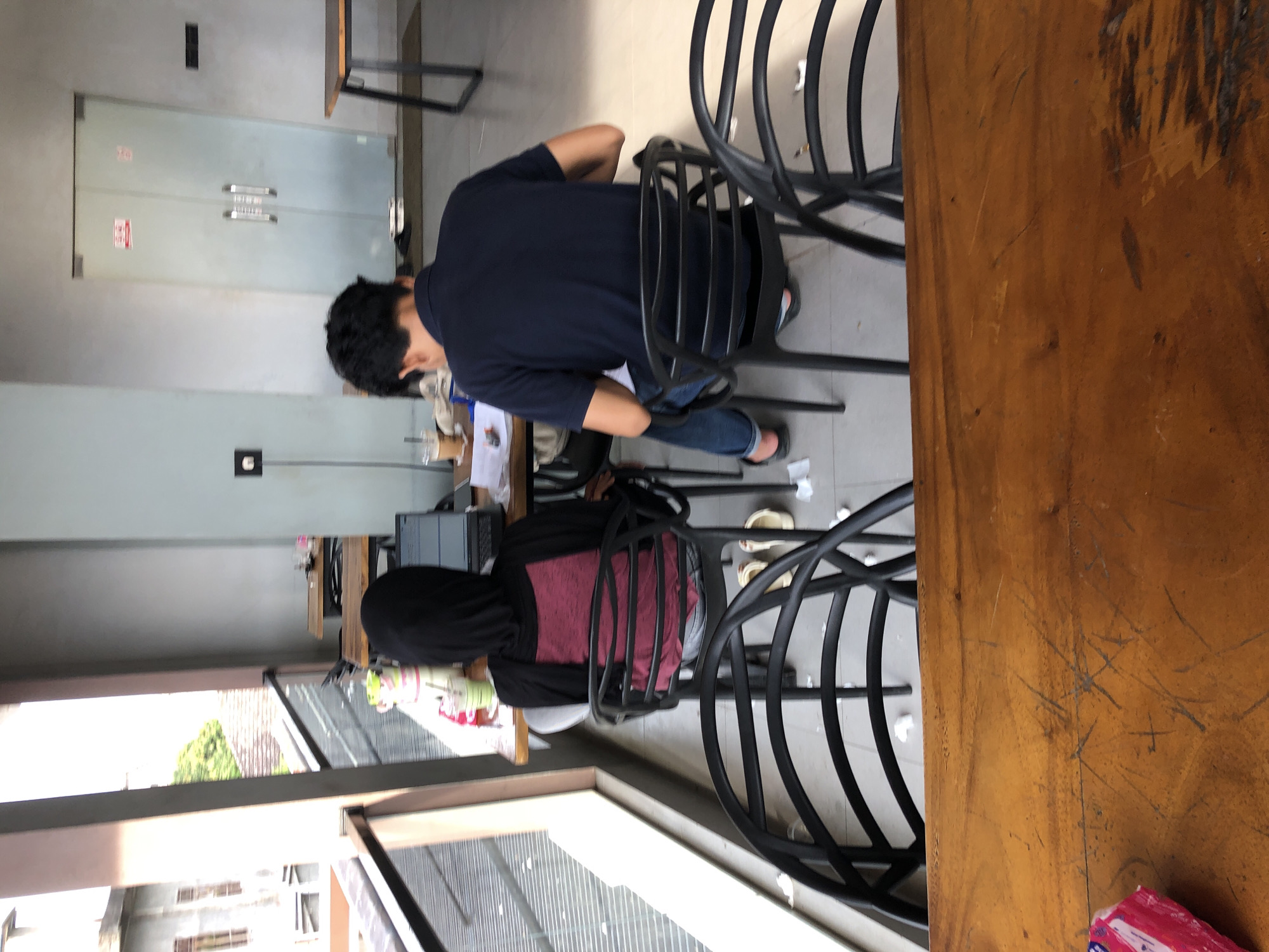 In Geulumpangsulu Timu, Indonesia, two individuals are seated at tables in an outdoor cafe or communal workspace. On the left, a person wearing a black hijab and a maroon top is facing away from the camera, looking at a laptop on a table. Next to the laptop are a drink with a straw and other miscellaneous items. To the right, a person wearing a dark blue polo shirt and blue jeans is also seated, facing away from the camera and looking down at a table, possibly working or studying. They are seated at a separate table from the first individual. The environment appears to be an open-air space with tables and chairs, with a building visible in the background and a balcony railing.