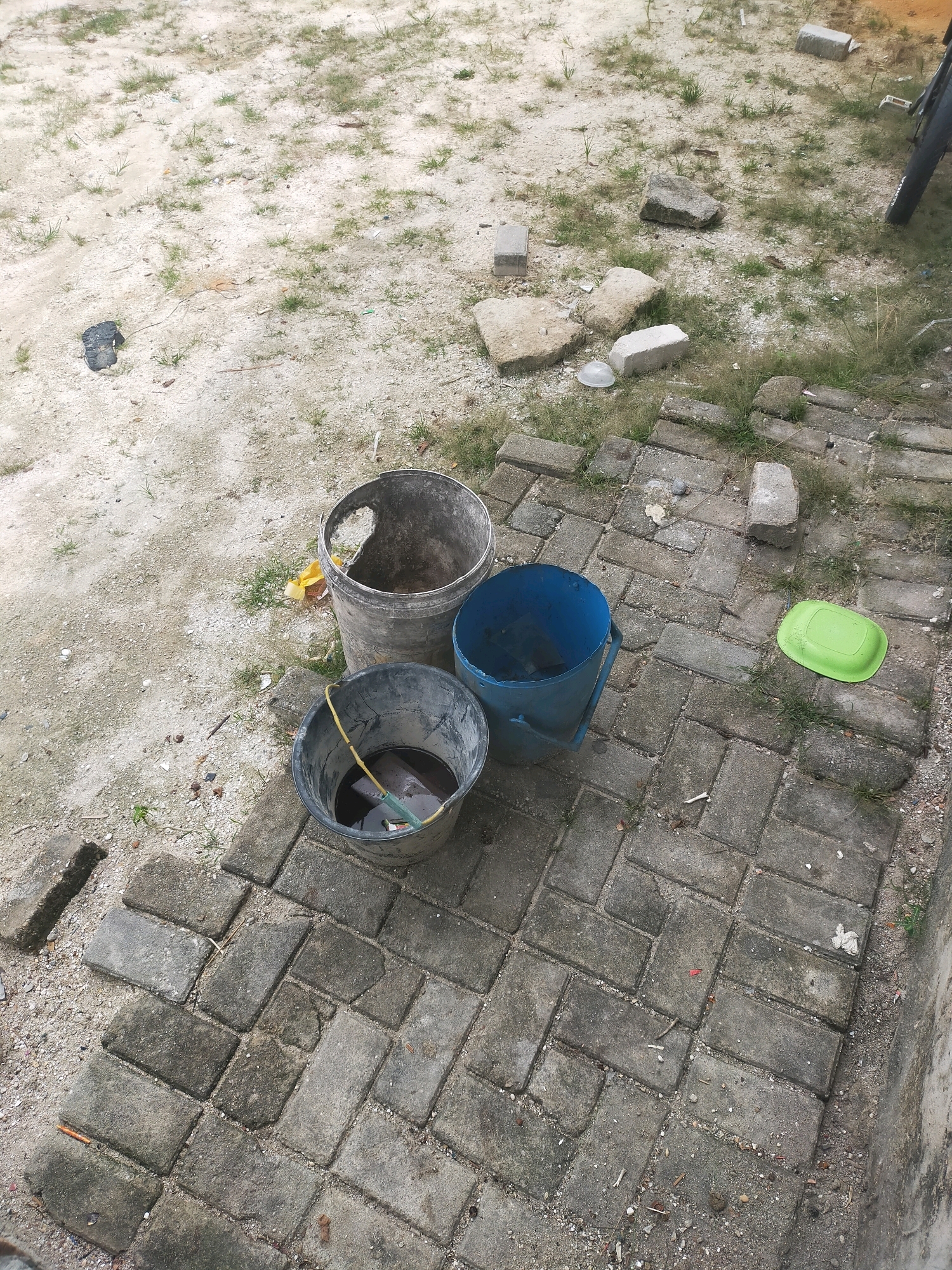 Three buckets and a green lid are on the ground in Pekanbaru, Indonesia. The largest bucket is silver and partially filled with debris. Next to it is a blue bucket, empty except for some dirt. The third bucket is black and filled with dark liquid, with a yellow handle protruding from it. The ground is a mix of dirt and gray paving stones. The scene appears to be outdoors, possibly a construction site or a backyard, with some scattered rocks and dry grass.