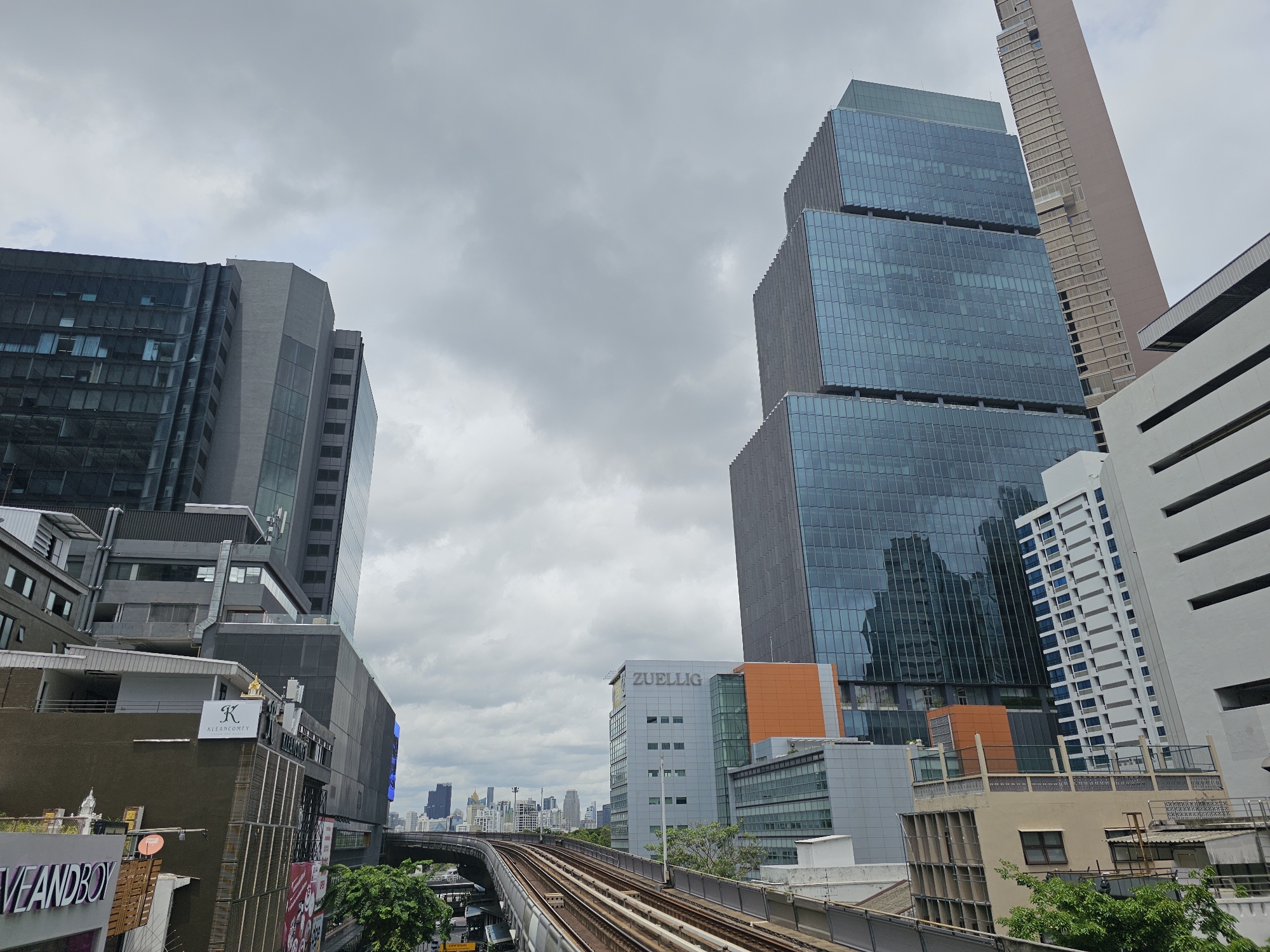 The image shows a view of the city of Bangkok, Thailand, dominated by modern high-rise buildings and an elevated railway track. The sky is overcast, with dense grey clouds. In the foreground, an elevated train track curves to the right. To the left of the tracks, a multi-story building with dark facades and some exposed balconies is visible. Further left, a taller, dark-glass skyscraper with multiple sections juts upwards. To the right of the train tracks and slightly further back, several contemporary buildings are present. One prominent building has a stepped design with dark blue glass facades. Another building, labeled "ZUELLIG" in large letters, features a lighter facade with orange accents. In the distance, a dense urban skyline with numerous skyscrapers can be seen. A tall, slender, light-brown skyscraper is visible behind the blue-glass building. The scene depicts a busy urban environment with a mix of architectural styles. There are no people or discernible actions visible in the image.
