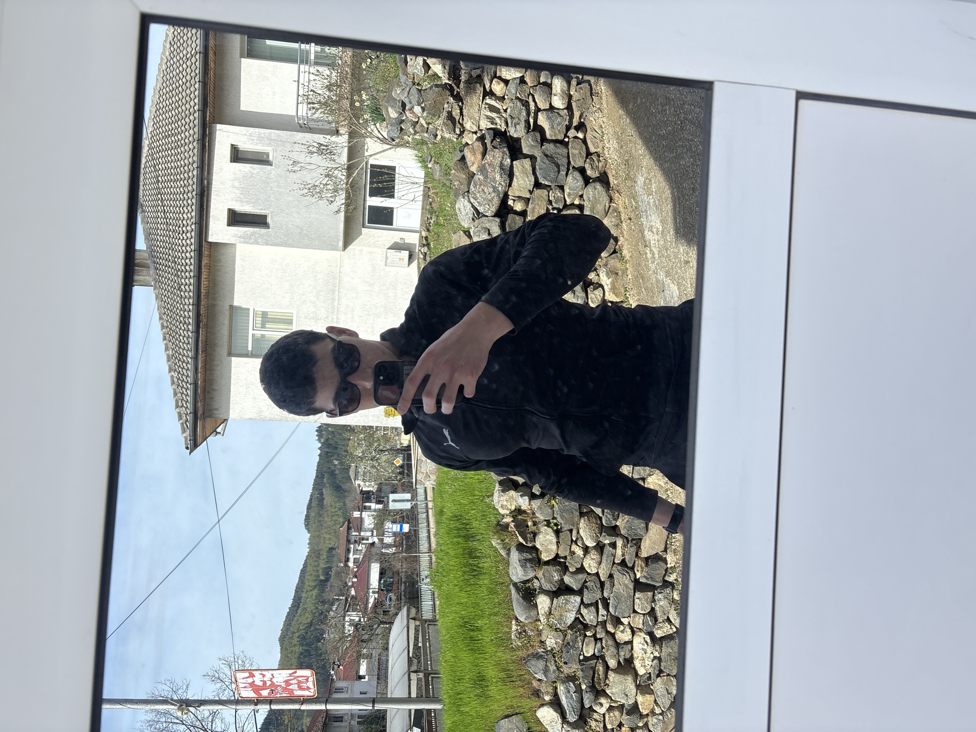 The image displays a selfie reflection of an individual in a window or glass door, set in Zlatograd, Bulgaria. The person is wearing dark sunglasses and a black hooded jacket, which features a white Puma logo on the left chest. They are holding a black smartphone to capture the reflection.
The outdoor scene, viewed through the window, depicts a bright and sunny day with a clear blue sky. Immediately outside, a rugged stone wall creates a natural barrier. Beyond this, a strip of bright green grass runs alongside what appears to be a road or pathway. The landscape features several houses with light-colored walls and distinct tiled roofs, interspersed with trees. In the background, lush green mountains rise under the clear sky, with power lines stretching across them.
Visible architectural elements include a prominent light-colored house with multiple rectangular windows. Two road signs are discernible: a red rectangular sign with some white, largely illegible text, and a smaller white sign with blue text, both situated near utility poles. The scene conveys a tranquil, daylight setting within a town nestled amidst a mountainous environment.