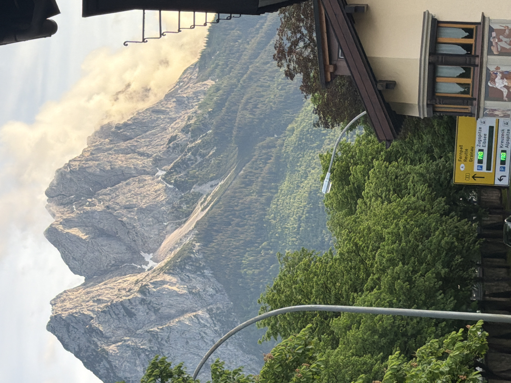This vertically oriented image (originally landscape, rotated) captures a majestic alpine scene with elements of a traditional town. In the background, a massive, rugged grey mountain range dominates the left side, exhibiting steep, rocky faces and some lingering patches of snow or ice. Wispy, warm-toned clouds partially obscure the mountain peaks, suggesting either early morning or late afternoon light. Lush, dense green forests cover the lower slopes of the mountain and extend into the midground, indicating a rich natural environment.
In the foreground and right side, parts of a building with traditional architecture are visible. It features a dark brown sloping roof, light-colored walls, and ornate wooden window frames. Below the windows, there are colorful murals depicting figures, possibly scenes of local life or history. A decorative black metal railing or ironwork is seen at the top edge of the building. A grey, curved metal pole, likely a streetlight arm or signage support, stretches across the lower-center of the frame, partially obscured by green foliage.
Attached to a pole on the right, a yellow directional sign with black text is prominent. The top section, with an arrow pointing straight ahead, lists destinations: "Fernpaß", "Reutte", "Grainau", and indicates "P Zugspitze" and "Elbsee" with a "FREE" symbol, likely for parking. The bottom section, with an arrow pointing to the right, shows "Kreuzeck" and "Alpspitz" also with a "FREE" parking symbol.
The scene depicts a peaceful, scenic view, likely from a village or town street, overlooking the mountains. The lighting is soft and natural, indicating good weather. No people are directly visible, but the presence of the building and signage suggests a populated area serving as a base for exploring the alpine landscape.