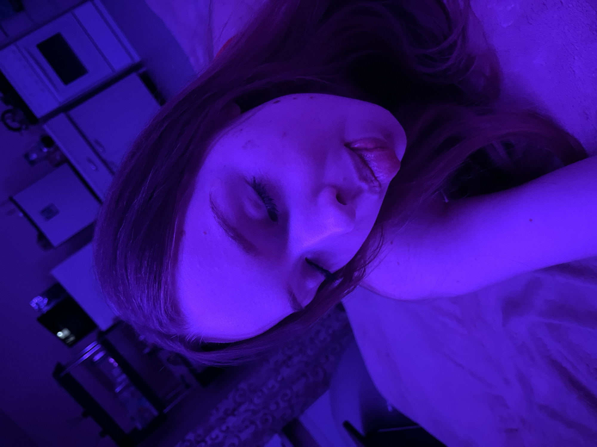 A close-up, rotated image depicts a person with light skin lying down. The person's face is angled towards the viewer with their eyes closed and mouth slightly open. Long, red hair surrounds the face. The lighting is predominantly blue, casting a strong hue over the subject and the background. In the background, there is a blurry view of what appears to be furniture, including cabinets and a television set. The location and city of Chaikovsky, Russia, cannot be determined.
