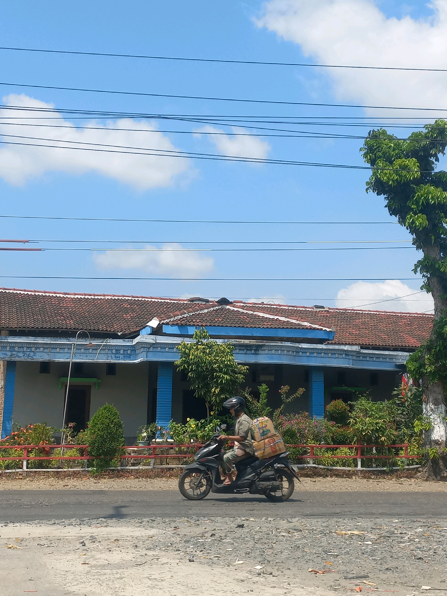 A man wearing a helmet and a light-colored shirt and pants rides a black motorcycle on a paved road. He is carrying two large cardboard boxes strapped to the back of the motorcycle. The motorcycle is in motion, captured mid-ride. Behind the motorcycle, there is a residential building with a tiled roof and blue trim around the windows and columns. A red and white fence runs in front of the building, with various plants and trees behind it. Power lines are visible in the bright blue sky above, with some scattered white clouds. The ground in the foreground is rough and appears to be a mixture of gravel and dirt. This scene is located on a road in Baturetno, Indonesia.