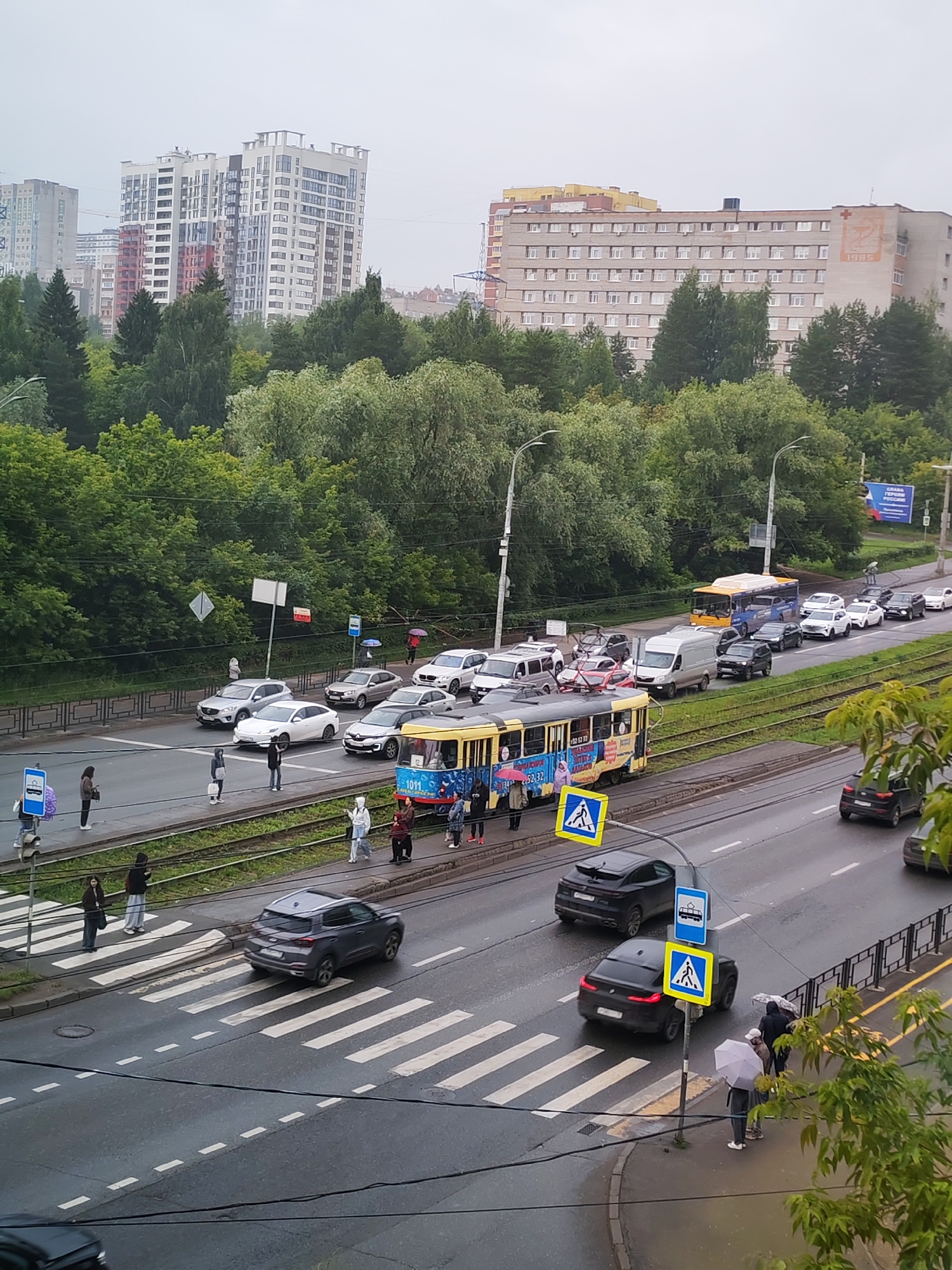 The image depicts a street scene in Izhevsk, Russia, on an overcast day, likely in the afternoon. The weather appears to be rainy or recently rained, as the road surface is wet and several individuals are holding umbrellas. The setting is urban, with a road running horizontally across the frame, flanked by trees and a tram line. In the background, modern apartment buildings and older institutional buildings are visible.
The main activity is traffic flow, with a considerable number of cars and a yellow tram stuck in traffic. The tram is decorated with colorful advertisements. Pedestrians are present at a crosswalk, waiting to cross the street. One person is walking their dog. The mood seems neutral, with the focus on the everyday urban environment.
Notable details include the various vehicles: cars of different models and colors, a blue bus, and the prominent yellow tram. Traffic signs, including pedestrian crossing signs with yellow backgrounds and blue signs with images, are visible. The buildings in the background suggest a developed urban area. Some text is visible on the tram, including numbers "52-52" and "1011", and possibly some advertising slogans. The building on the right side of the background has the number "1985" and a red cross symbol.