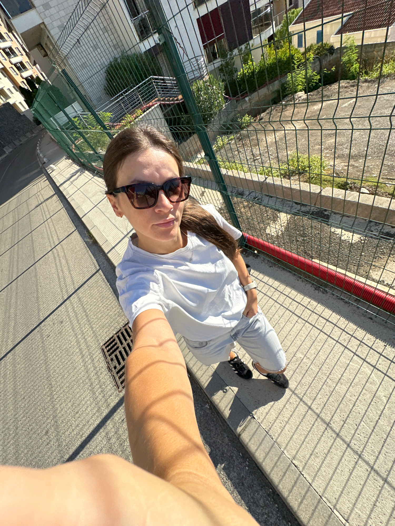 This media file shows a young woman taking a selfie outdoors in what appears to be a residential area. She is wearing a white t-shirt, light-colored shorts, and black sneakers. Her hair is dark brown and pulled back into a ponytail. She is also wearing sunglasses with dark frames. In the background, there is a metal fence, buildings, and greenery. The sun is shining brightly, casting strong shadows from the fence onto the concrete walkway. A red tube runs along the base of the fence. The scene suggests it is daytime, likely a sunny day. The woman's expression is neutral, as she is looking towards the camera. The overall impression is of a casual, everyday moment. The location context provided is Boreti, Montenegro.