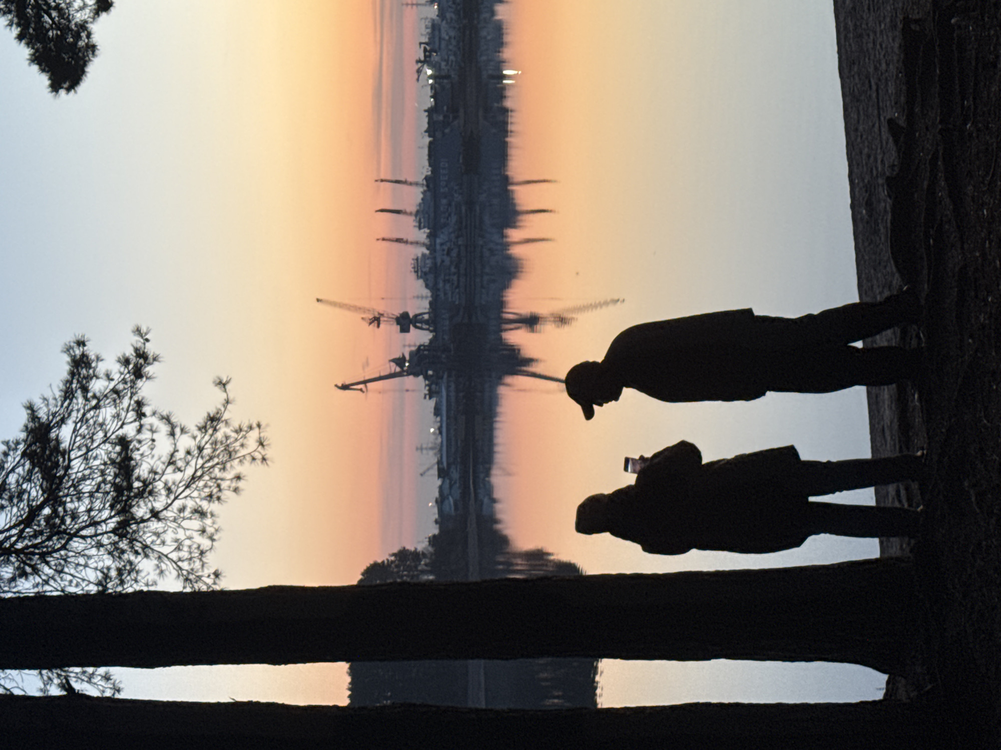 The image depicts a serene waterfront scene captured during either sunrise or sunset in Chaikovsky, Russia. Two individuals are silhouetted against the vibrant sky on the right side of the frame, standing beside a calm body of water, likely the Kama River or a related reservoir.
One figure, appearing to be a man, wears a cap and stands slightly behind another person who holds a smartphone up, seemingly capturing a photograph or video of the picturesque view. Their forms are sharply contrasted against the luminous sky.
The sky itself transitions from a soft pale blue on the left to a warm gradient of orange and pink on the right, characteristic of the golden hour. Across the tranquil water, a distant cityscape or industrial complex is perfectly reflected. Visible in these reflections are numerous silhouettes of buildings, several prominent cranes suggesting a port or industrial zone, and possibly moored vessels. The stillness of the water creates an almost mirror-like surface, doubling the visual elements of the distant shore and the sky.
In the foreground, dark silhouettes of tree branches frame the left side and top-left corner of the image. A dark, indistinct horizontal barrier or bank runs across the bottom of the frame, separating the viewers from the water. The overall impression is one of peaceful observation, with the strong silhouettes and warm, reflective light dominating the composition. There is no visible text in the image.