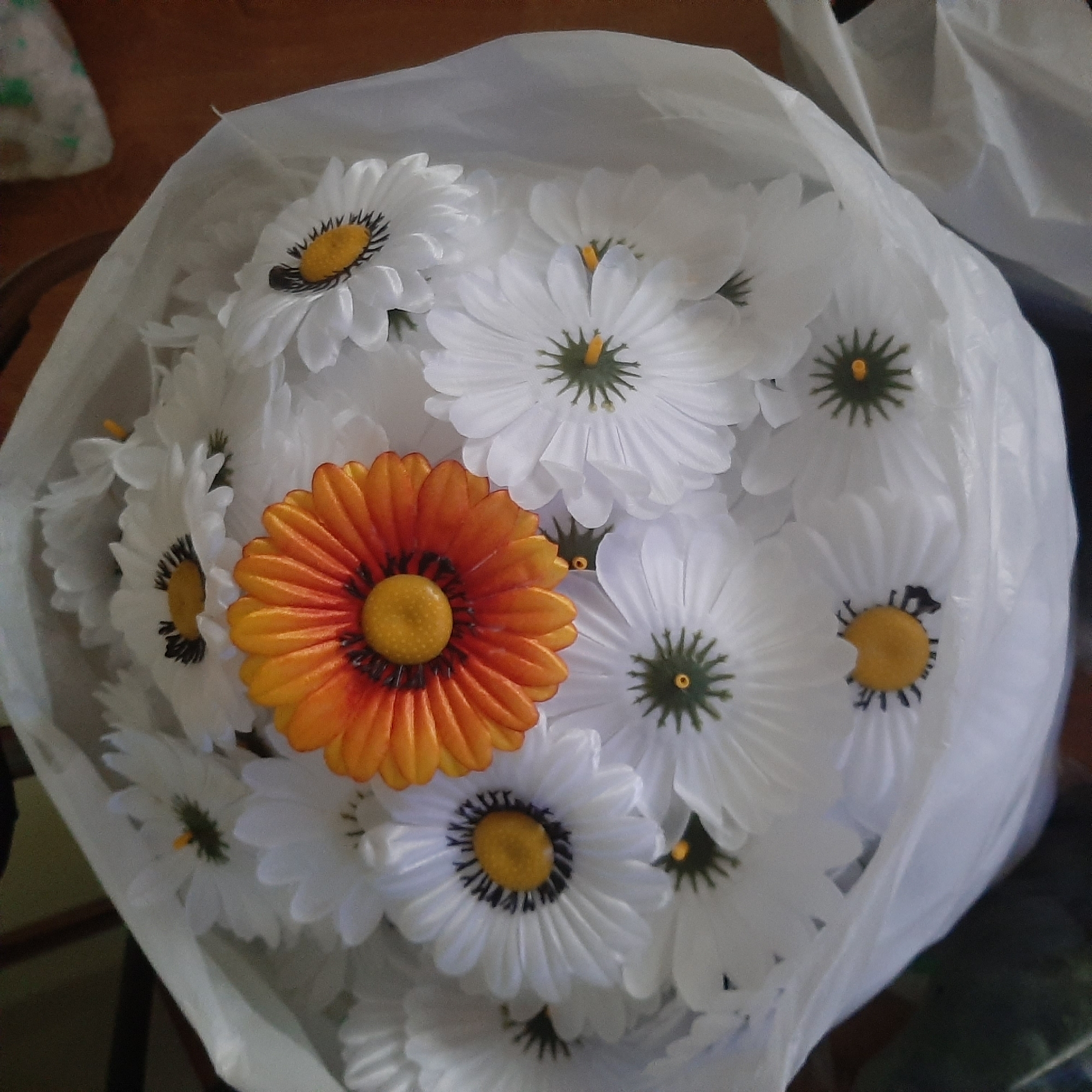 In the image, a bouquet of artificial daisies is presented. The majority of the flowers are white with yellow centers, featuring black accents around the pistils. One prominent flower in the center is a vibrant orange, also with a yellow center and radiating orange petals. The flowers are arranged within a white plastic bag, which is partially obscuring the bouquet. The background suggests an indoor setting, with a wooden surface visible in the upper right and left portions of the image. The lighting is somewhat dim, and the focus is on the flowers themselves. The location is unknown, as the city and state are not specified.