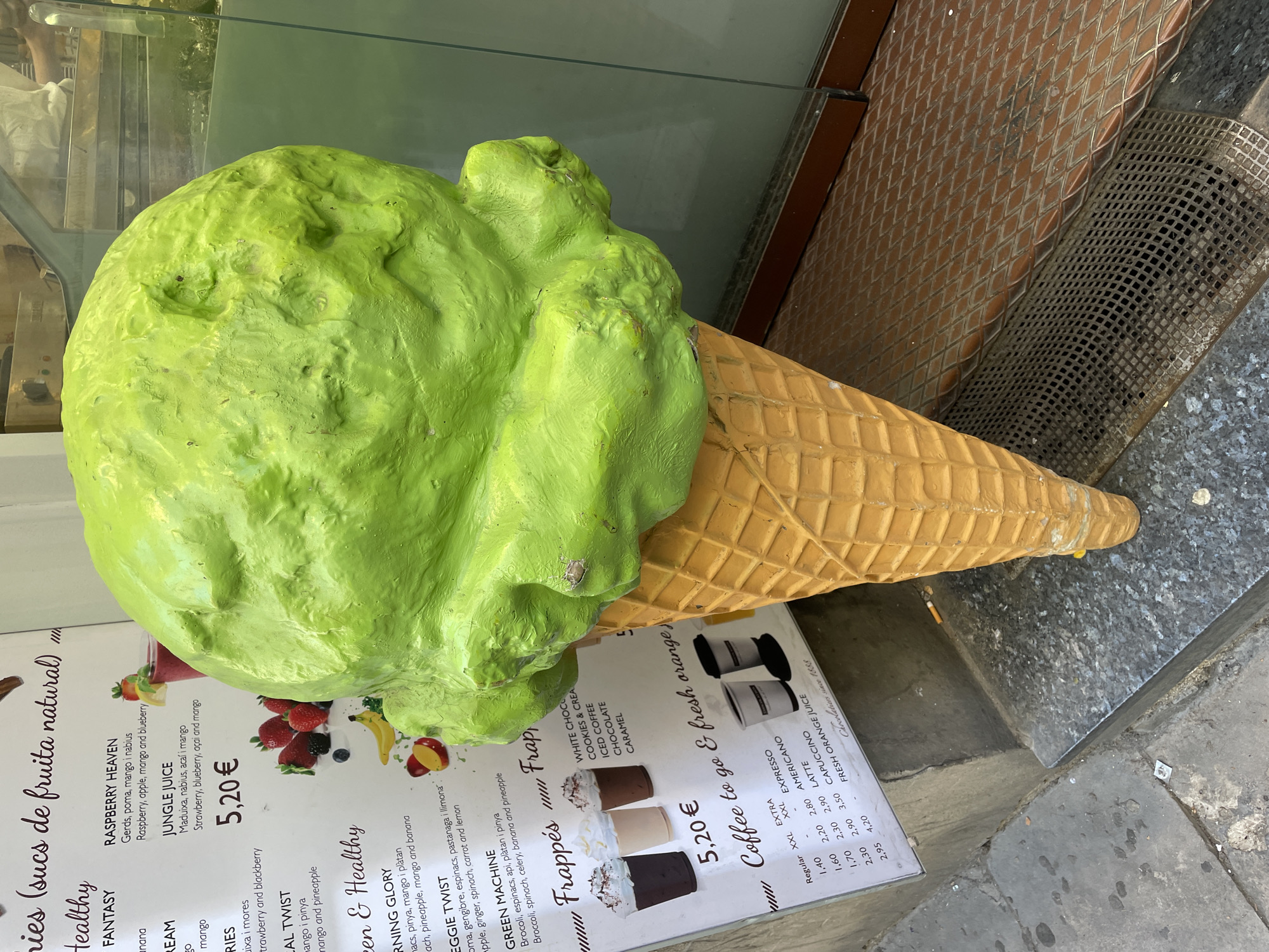 A large, bright green, oversized ice cream cone sculpture serves as a prominent storefront display, positioned on the ground next to a building. The cone itself is a light brown, waffle-patterned replica, while the "scoop" of ice cream is a vibrant lime green, with an irregular, somewhat melted or soft-serve texture.
Behind and partially obscured by the ice cream cone is a menu board, leaning against the building. The menu advertises "sucs de fruita natural" (natural fruit juices), with sections like "Healthy Fantasy" and "Green & Healthy." Specific juice blends mentioned include "RASPBERRY HEAVEN" (Raspberry, apple, mango and blueberry), "JUNGLE JUICE" (Strawberry, blueberry, açai and mango), and "GREEN MACHINE" (Broccoli, spinach, celery, banana and pineapple), all priced at 5,20 €. The menu also lists "Frappés" for 5,20 €, featuring flavors such as "WHITE CHOC," "COOKIES & CREAM," "ICED COFFEE," "CHOCOLATE," and "CARAMEL." Another section offers "Coffee to go & fresh orange," with prices for "EXPRESSO," "AMERICANO," "LATTE," and "CAPUCCINO" in various sizes (Regular, XXL, EXTRA XXL) ranging from 1,40 € to 4,20 €. The phrase "Chocolatiers since 1868" is also visible.
The setting appears to be an urban street or sidewalk, under bright, possibly daytime, lighting. Reflections are visible on the glass surface behind the ice cream. The ground consists of grey stone or concrete slabs, alongside textured metal grates. A discarded cigarette butt lies on the ground near the base of the cone. The overall scene indicates a cafe or juice bar aiming to attract passersby with its colorful, larger-than-life prop and diverse menu offerings.