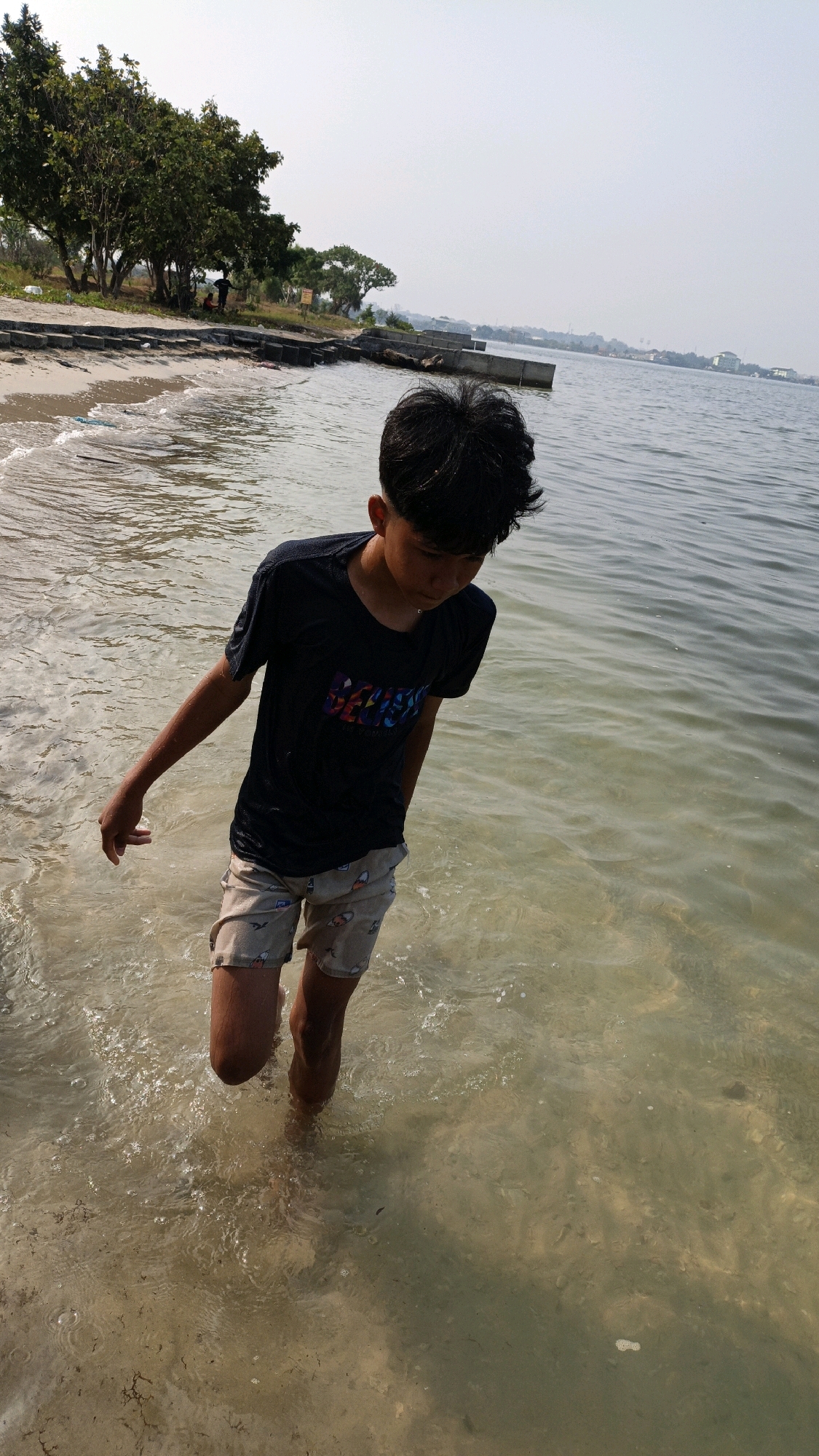 A young boy with dark hair and a dark t-shirt walks through shallow, clear water near a sandy beach. He is wearing patterned shorts and looking down at the water. Trees and structures are visible in the background along the shoreline, with buildings and distant landmasses in the far distance across the water. The sky is clear and overcast. This scene is consistent with a coastal area in Cikal, Indonesia.