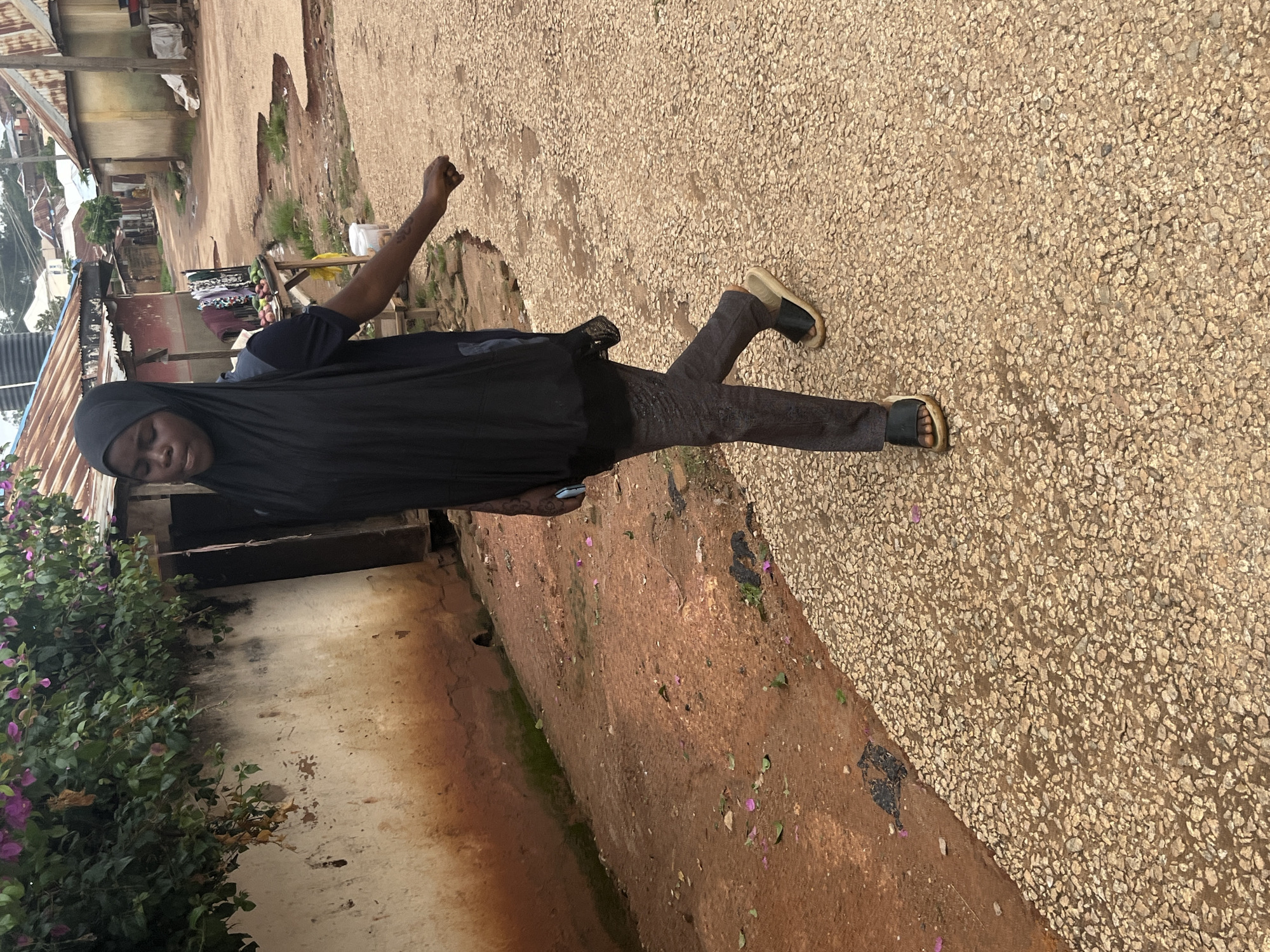 A person wearing a dark hijab and black clothing stands in the middle of a pathway. The pathway is made of small, light-colored stones. The person's left arm is raised with a clenched fist. They are wearing dark pants and black sandals with light-colored soles. To the left of the pathway is a wall covered in green foliage with purple flowers. Behind the person, the pathway continues, with buildings visible on either side. One building appears to have a corrugated metal roof. The scene suggests an outdoor, possibly urban or semi-urban environment in Jos, Nigeria. The lighting suggests it is daytime.