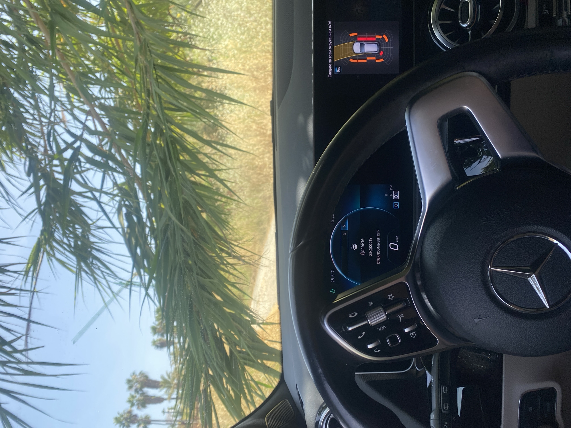 The image shows the interior of a car, specifically the dashboard and steering wheel, as viewed from the driver's seat. Outside the car, through the windshield, a sunny, arid landscape with palm trees is visible, suggesting a warm climate. The scene appears to be in Bel Air, Spain.
The dashboard displays several indicators. On the right side, a parking assist system is active, showing a graphical representation of the car and its surroundings with proximity warnings. On the left side of the dashboard, a digital display shows the current temperature as 28.5°C and a message in Russian that translates to "Top up windshield washer fluid". Below this, the speed is indicated as 0 km/h, suggesting the car is stationary.
The steering wheel is a prominent object, featuring the Mercedes-Benz logo in the center. Various controls are visible on the steering wheel, including buttons for media playback, phone calls, and cruise control. The overall impression is that of a car parked in a sunny, possibly rural or suburban, location in Spain on a clear day. No people are visible in the image.