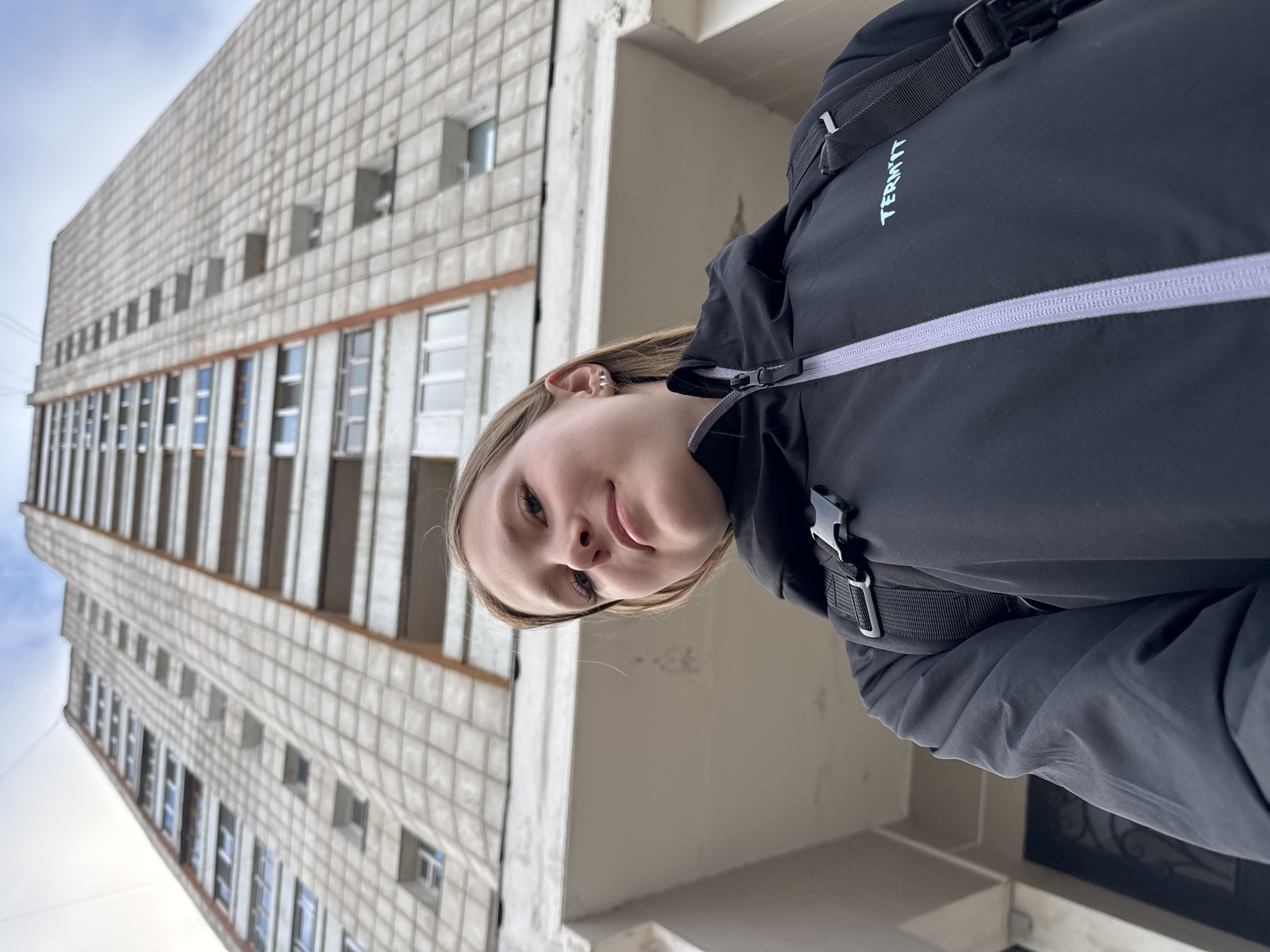 The image, taken with a low-angle perspective and rotated 90 degrees, features a young woman posing for a selfie in front of a tall, utilitarian-style building in Chaikovsky, Russia.
The woman, with light-colored hair, is positioned in the foreground, occupying the lower right portion of the frame. She looks directly towards the camera with a neutral to slightly smiling expression. Notable details include a three-stud earring on her visible left ear. She is wearing a black jacket, which appears to be a durable outdoor garment, possibly a windbreaker or light puffer. A prominent light purple/grey zipper crosses the chest area of her jacket, and a light blue/teal logo spelling "TERMI'T" is visible on the upper left chest. Part of a black strap and buckle, likely from a backpack or jacket feature, is visible near her right shoulder.
The background is dominated by a large, multi-story building constructed with concrete panels, characteristic of post-Soviet architecture. Numerous rectangular windows, some open, are visible across its facade, reflecting parts of a cloudy sky. The building appears weathered but structurally sound, suggesting a residential or administrative block. The sky above is partially cloudy, indicating an overcast or cool daytime setting. The overall scene conveys an urban environment, likely in the provided location of Chaikovsky, Russia, given the architectural style. The activity depicted is a self-portrait or selfie outdoors.