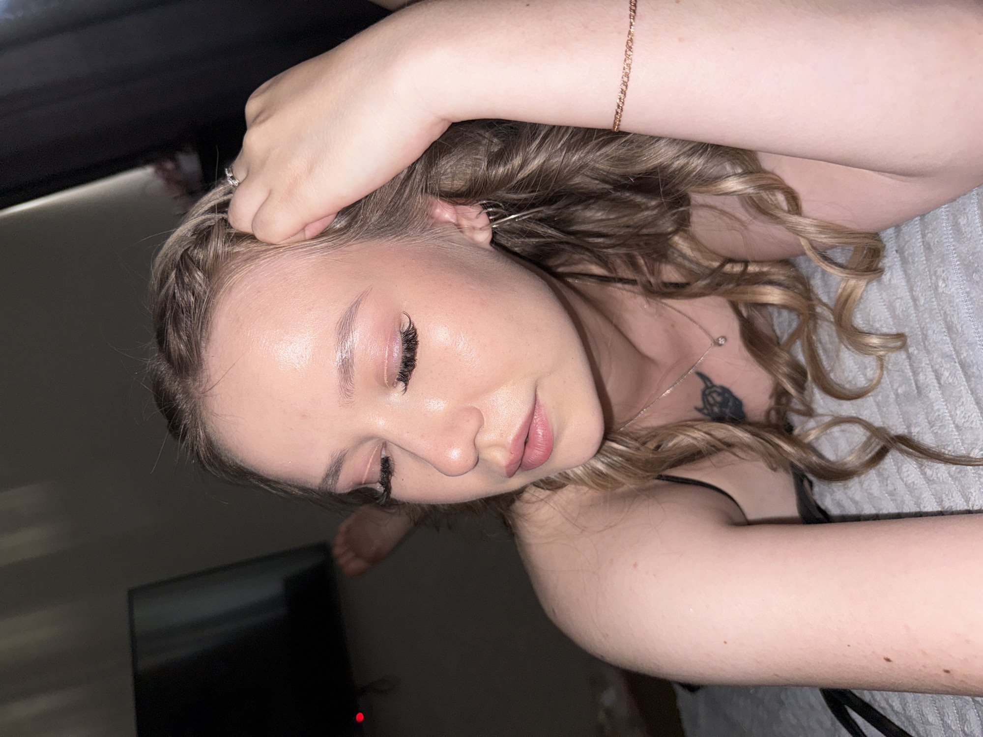 This is a medium close-up shot of a young Caucasian woman lying on her side on a textured, light-colored surface, possibly a bed. She has fair skin, light brown hair with blonde highlights styled in loose waves and a braid, and is wearing subtle makeup including pink lipstick and prominent false eyelashes. Her eyes are closed, and her head is tilted down, with her right arm resting above her head and her left arm bent across her chest. A delicate silver necklace with a small pendant is visible, as well as a bracelet on her right wrist. She also appears to have a small tattoo on her upper chest. In the background, to the left, a dark television screen is visible with a red indicator light. The overall lighting is dim, with some light casting shadows on the wall behind her. The image does not contain any elements that can be used to determine its location within the city of Chaikovsky, Russia.
