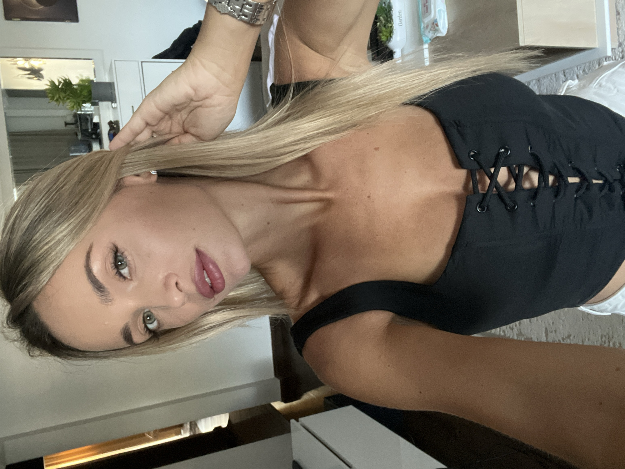 A woman with blonde hair and green eyes, wearing a black corset-style top, is taking a selfie in a room. She is holding her hair back with her right hand, and a silver bracelet is visible on her left wrist. The room appears to be a bedroom or living area, with a dresser and mirror in the background. There is a plant on the dresser and some framed artwork on the wall. The lighting suggests it is daytime. The image does not contain any explicit information about the location being Sharm El Sheikh, Egypt.