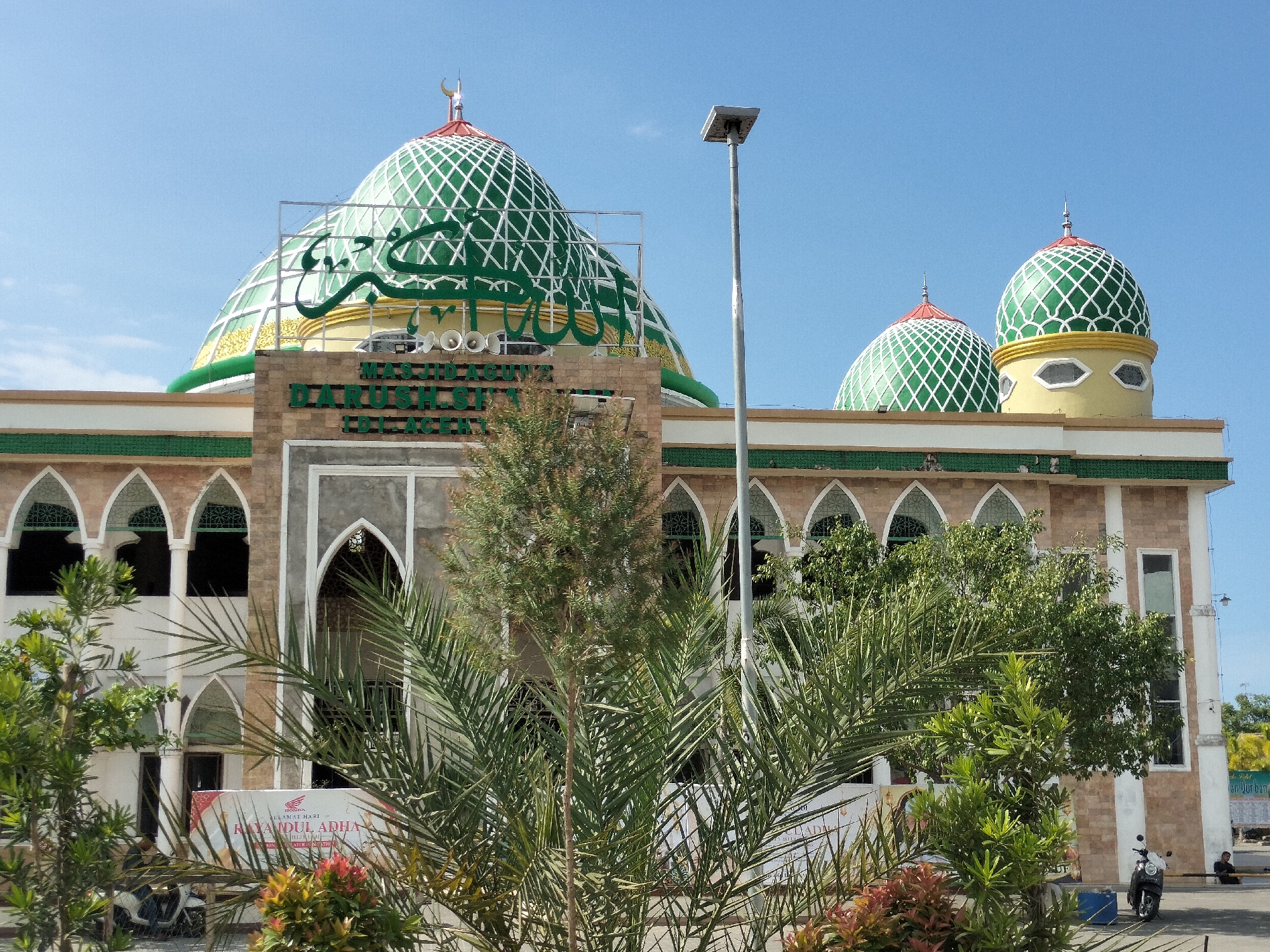 The image shows the Darussalam Mosque in Titibaro, Indonesia. The mosque features a large central dome and two smaller domes, all with green lattice patterns and crescent moon finials. The building's facade is constructed of tan brick with light-colored arched windows. A sign reading "MASJID AGUNG DARUSSALAM SGI ACEH" is prominently displayed above the entrance. Trees obscure a clear view of the lower portion of the mosque. To the left is a poster that says "Selamat Hari Raya Idul Adha", indicating it is a holiday celebration. A light pole rises vertically in the background. Parked motorcycles and people can be seen at the edges of the frame. The sky is clear and blue.