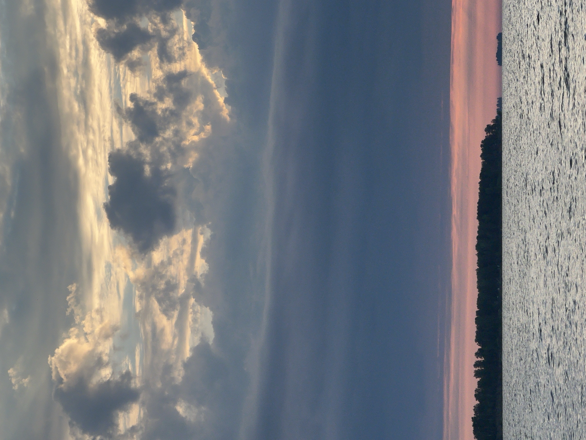 The image captures a serene natural landscape at dusk, likely near Borovoe, Russia. The scene is dominated by the vast expanse of a calm body of water in the foreground, reflecting the muted light. In the distance, a dark, silhouetted treeline marks the edge of the land, with hints of a soft pink and orange sunset peeking through the horizon. Above the treeline, the sky is a dramatic gradient of deep blues and greys, transitioning into lighter, warmer tones where the last rays of sunlight catch the edges of the clouds. The clouds themselves are varied in texture and density, some appearing wispy and translucent, while others are thicker and more opaque, creating a sense of depth and atmosphere. There are no people or man-made structures visible in the image, emphasizing the untouched beauty of the natural environment.