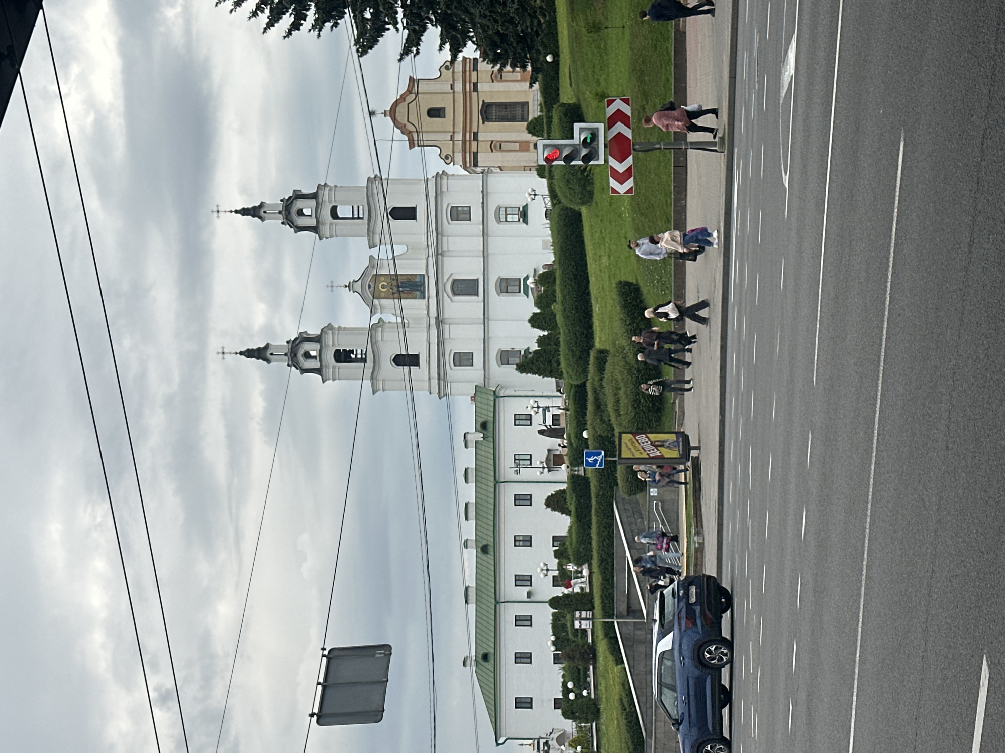 The image depicts a street scene in Minsk, Belarus, featuring a large white church with two prominent bell towers and a smaller adjacent yellow building. In the foreground, a blue car is parked on the left side of the frame, partially obscuring a set of stairs leading up a grassy embankment. Pedestrians are seen walking along the sidewalk in front of the church and buildings. Traffic lights are visible, with the top light illuminated red, indicating a stop. A large billboard with text and an image of a woman is also present on the right side of the sidewalk. The sky is overcast, suggesting a cloudy day. Overhead power lines are strung across the upper portion of the image. The overall scene appears to be a public area with a mix of architectural and urban elements, typical of a city setting.