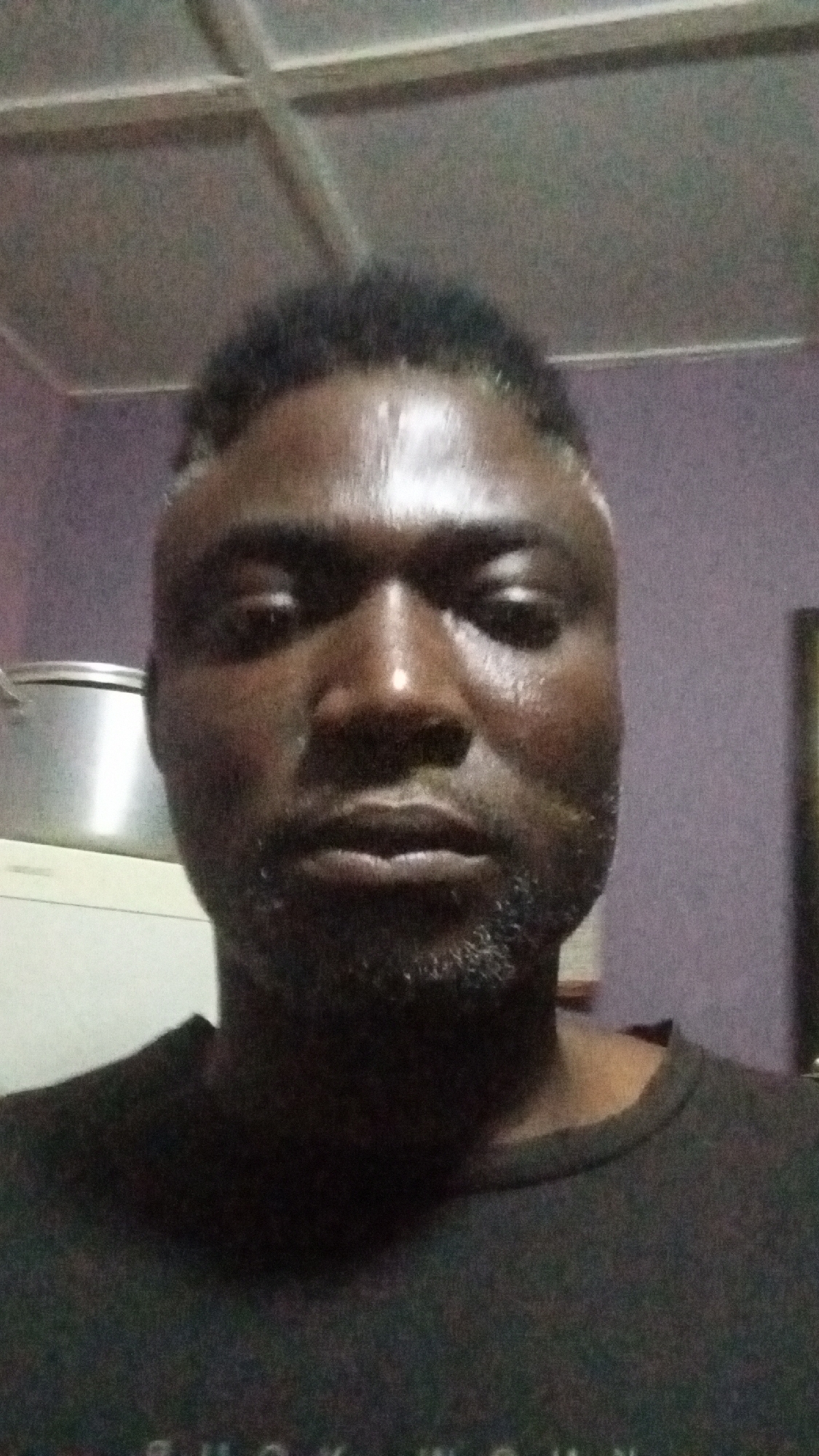 A medium indoor shot depicts a dark-skinned man with short, dark hair and a beard. His face appears slightly damp or sweaty. He is wearing a dark t-shirt. In the background, a metal pot is visible on a white appliance. The background wall appears to be purple. The ceiling appears to be made of light-colored panels. There is no information available to confirm if this photo was taken in Sabon Tasha, Nigeria.