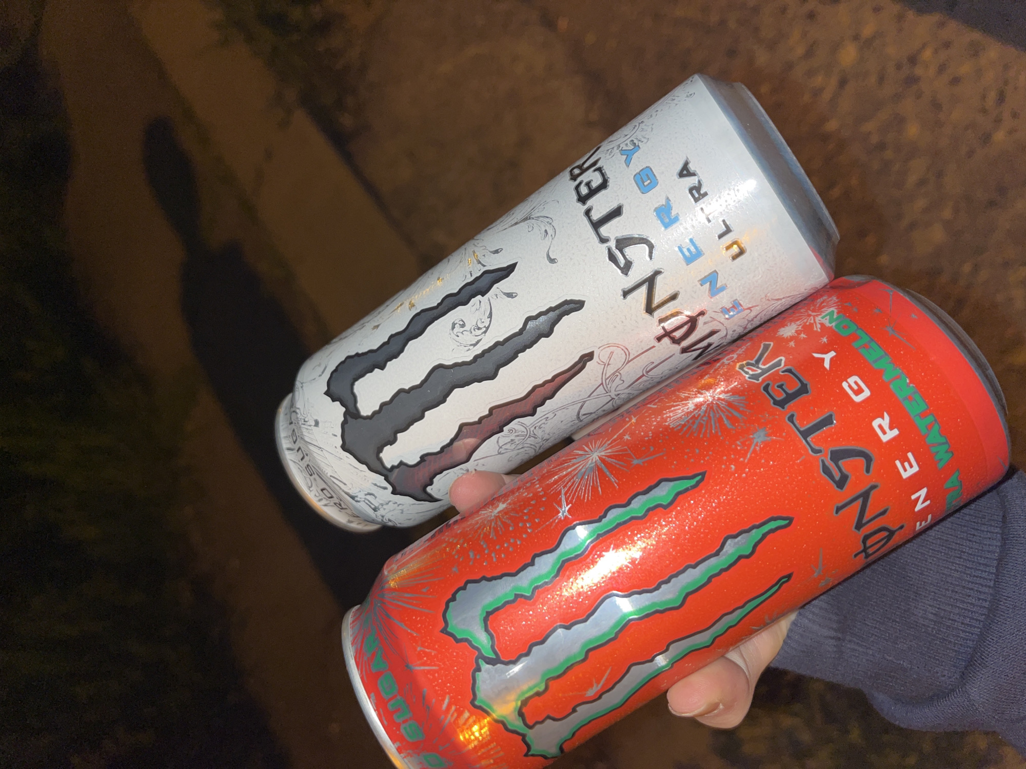 The image displays a close-up of a person's hand holding two Monster Energy drink cans, set against a dark, indistinct outdoor background. The hand holds one can prominently in the foreground, while the other is visible slightly behind it.
The top can is white, featuring a silver and grey Monster claw logo. Elaborate white and silver patterns adorn its surface. Visible text on this can includes "MONSTER ENERGY ULTRA ZERO SUGAR". The bottom can is a vibrant red, showcasing a silver and green Monster claw logo. Its design incorporates festive silver sparkle graphics, suggesting a celebratory or special edition. Text visible on this can identifies it as "MONSTER ENERGY ULTRA WATERMELON ZERO SUGAR". Both cans appear to have condensation, indicating they are cold.
The setting is outdoors, likely at night or in very low light, as evidenced by the dark background and a long, distinct shadow of a person cast on the ground to the left. The ground surface is dark and unidentifiable beyond being a paved or natural ground cover. The lighting suggests a single, artificial light source illuminating the cans and casting the shadow.
The activity depicted is simply the holding of the two energy drinks. No specific event or strong emotions are discernible. The provided location context is Sofia, Bulgaria.