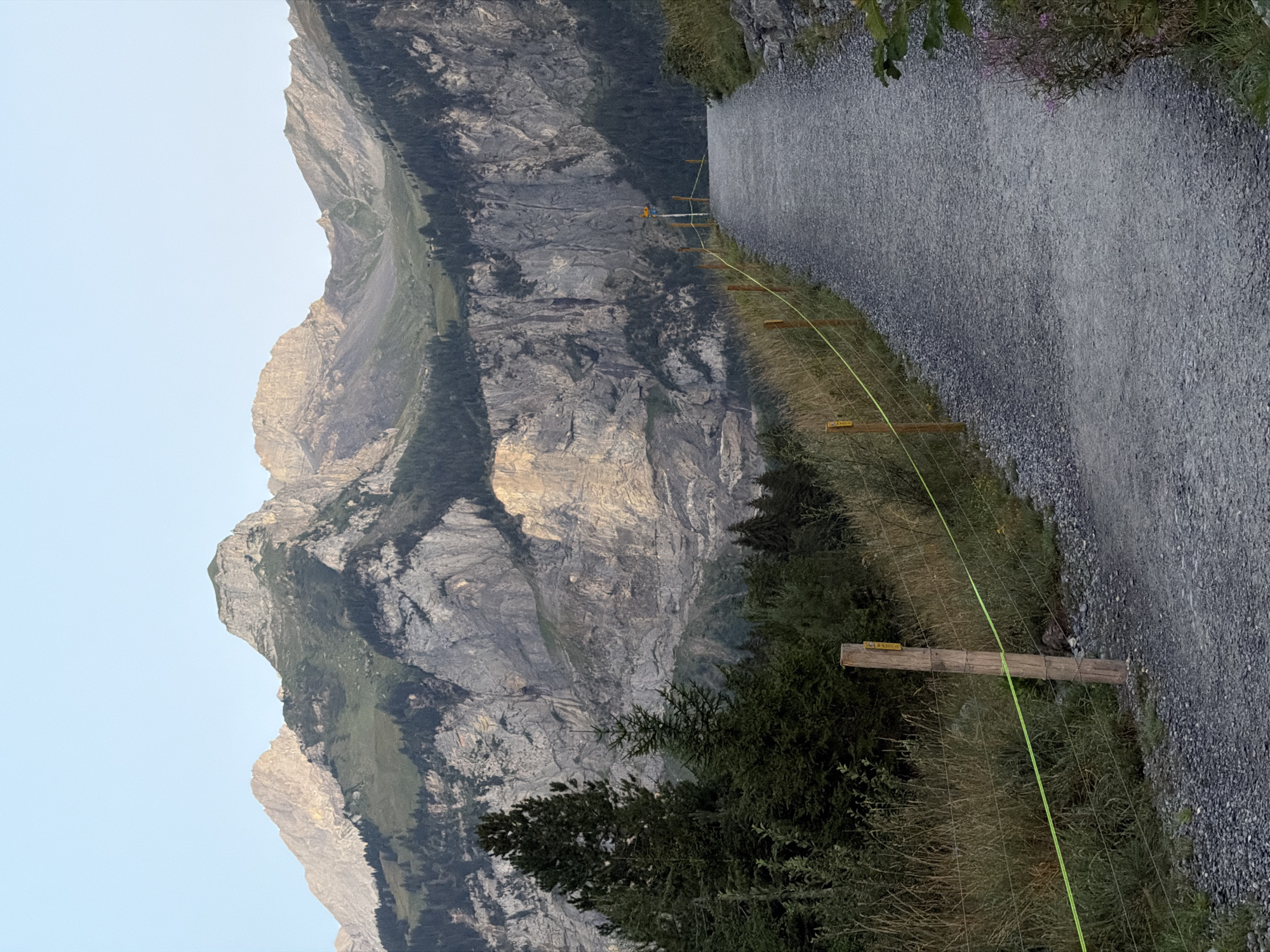 The image shows a gravel path leading uphill towards a majestic mountain range in Kandersteg, Switzerland. The path is bordered on the left by a wooden fence with bright yellow tape, separating it from a grassy embankment dotted with sparse trees. The mountains in the background are rugged and rocky, with patches of green vegetation and dark forests clinging to their slopes. The sky above is a clear, pale blue. The overall scene depicts a scenic and tranquil mountain landscape, likely a hiking trail, bathed in the soft light of early morning or late afternoon.