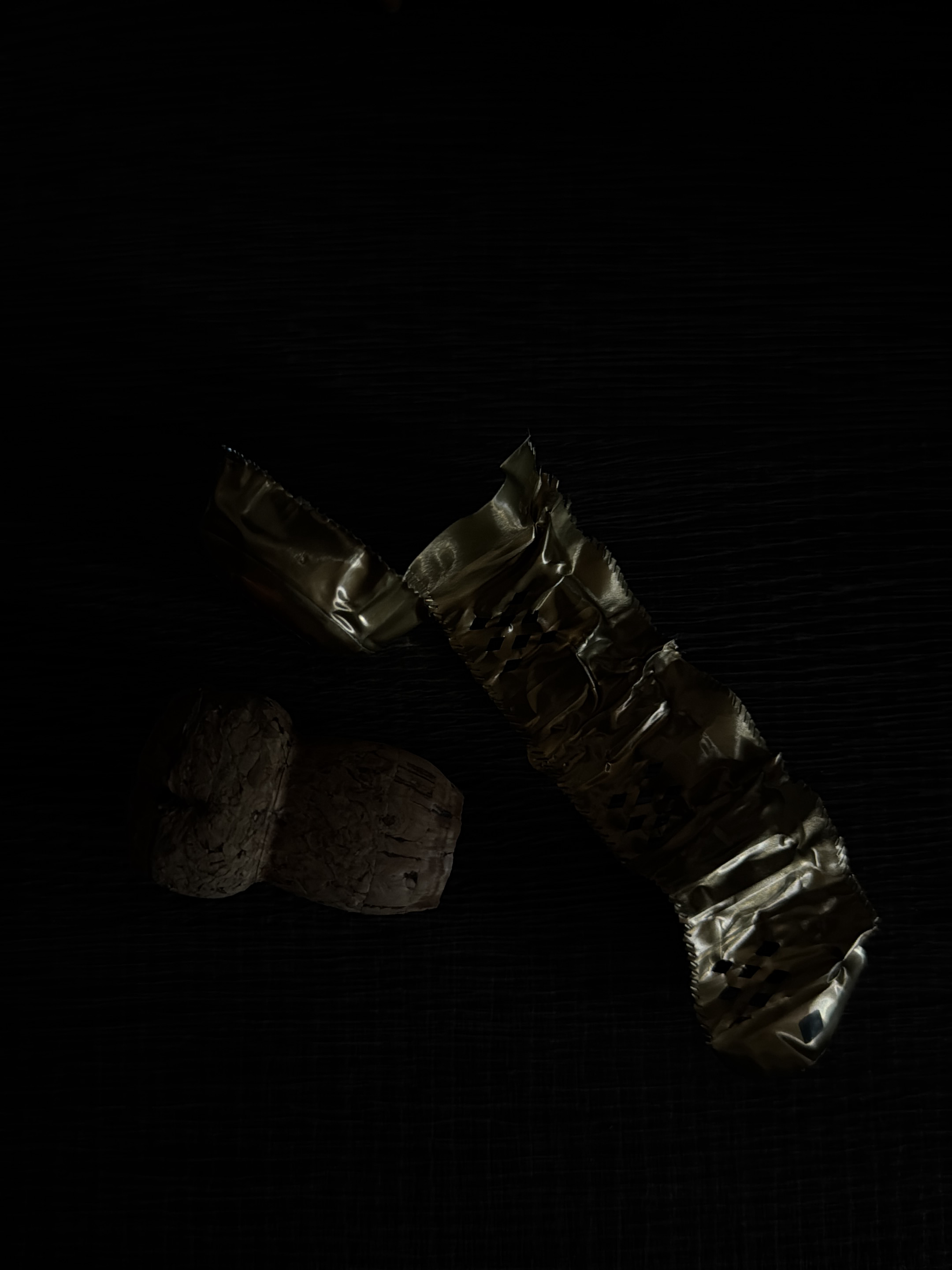 The image shows a champagne cork and a piece of foil on a dark, textured surface. The cork is on the left, appearing to be made of compressed wood. The foil is on the right, crumpled and reflective. The lighting is dramatic, casting deep shadows and highlighting the textures of the objects. The background is completely dark, with faint horizontal lines suggesting a wooden surface. The image provides no information about a specific location in Milan, Italy.