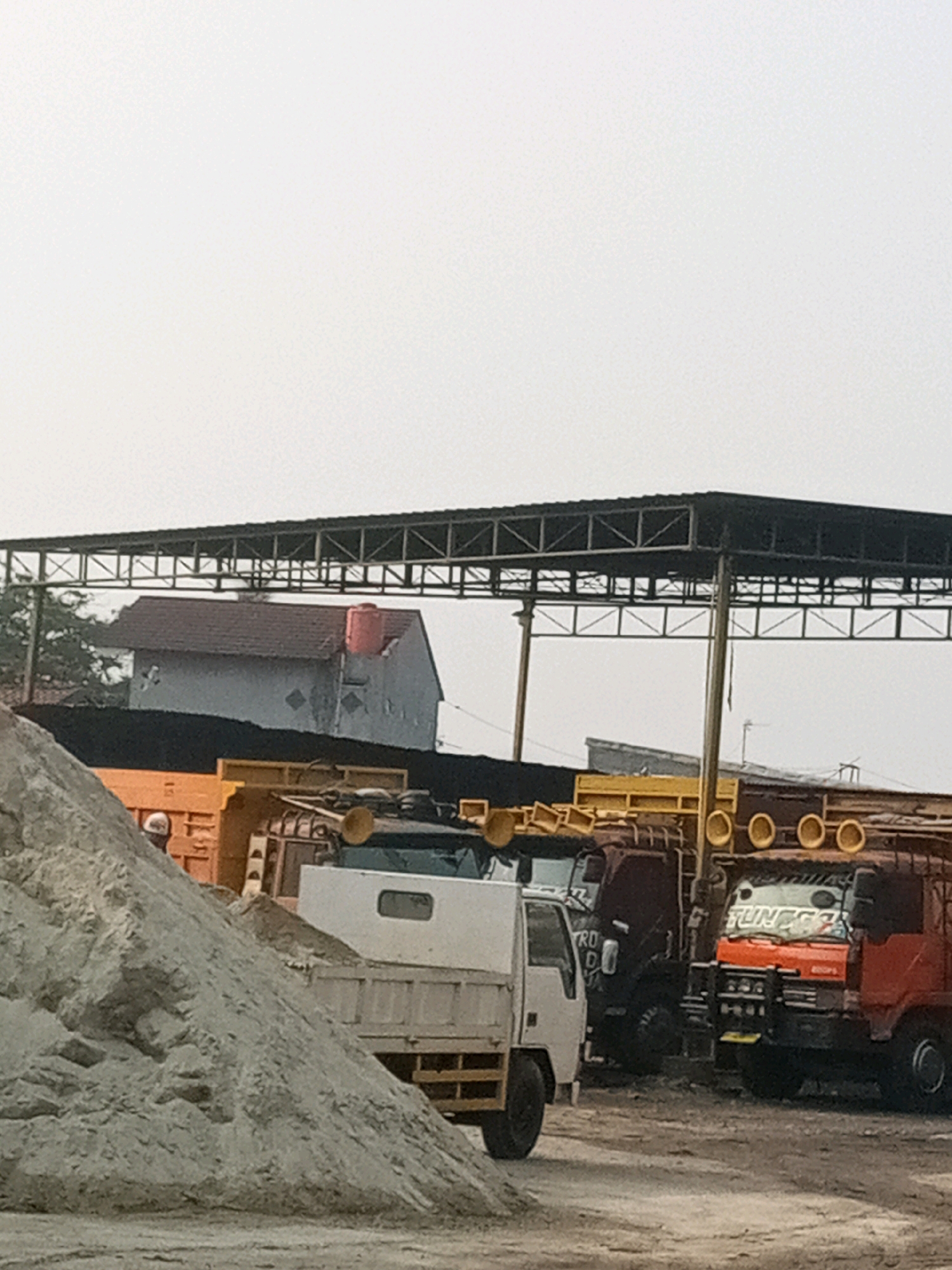 The image depicts an outdoor scene in Bekasi, Indonesia, likely a storage or construction yard. In the foreground, a large pile of light gray sand dominates the left side. Behind the sand pile, several trucks are parked under a large, dark metal canopy. One truck, partly visible on the far left, is orange and appears to be a dump truck. Next to it is a white flatbed truck. Further to the right, a dark colored truck is visible, and to its right, another dump truck with an orange cab and a black grill. The background features a simple, light-colored building with a red water tank on its roof, and some trees. The sky is overcast and pale. The scene is static, with no apparent activity or interaction.