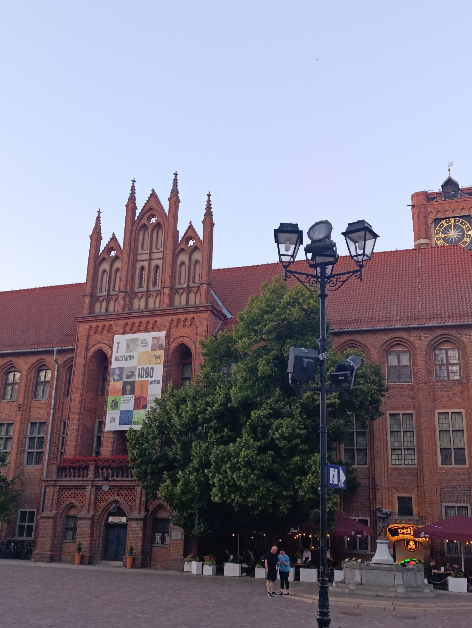 The image shows a view of the Old Town Hall in Toruń, Poland. The building is made of red brick and has Gothic architectural features, including pointed arches and spires. A large banner with text and images is displayed on the facade. A street lamp with multiple lights is prominent in the foreground, and a clock tower is visible on the right side of the building. Green trees are growing in front of the building, and outdoor seating with umbrellas is set up. Two people, a man and a woman, are standing near the street lamp, facing away from the camera. A fountain with a statue is also visible. The sky is clear and light blue.
