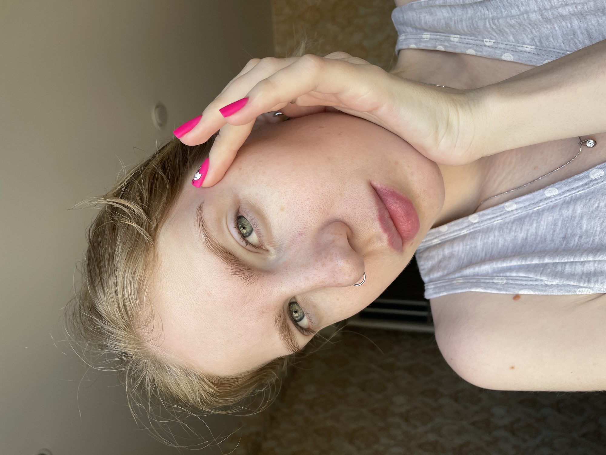 A fair-skinned woman with blonde hair and green eyes is photographed indoors. She is wearing a light grey t-shirt with white polka dots and a silver necklace. Her fingernails are painted bright pink, and one fingernail has a small white design on it. She has a nose ring on her left nostril. Her right hand is touching her forehead. The background appears to be a wall with a neutral color and a section of flooring visible at the bottom of the image with a textured pattern. The location in the city of Izhevsk, Russia, is not discernible from the image.