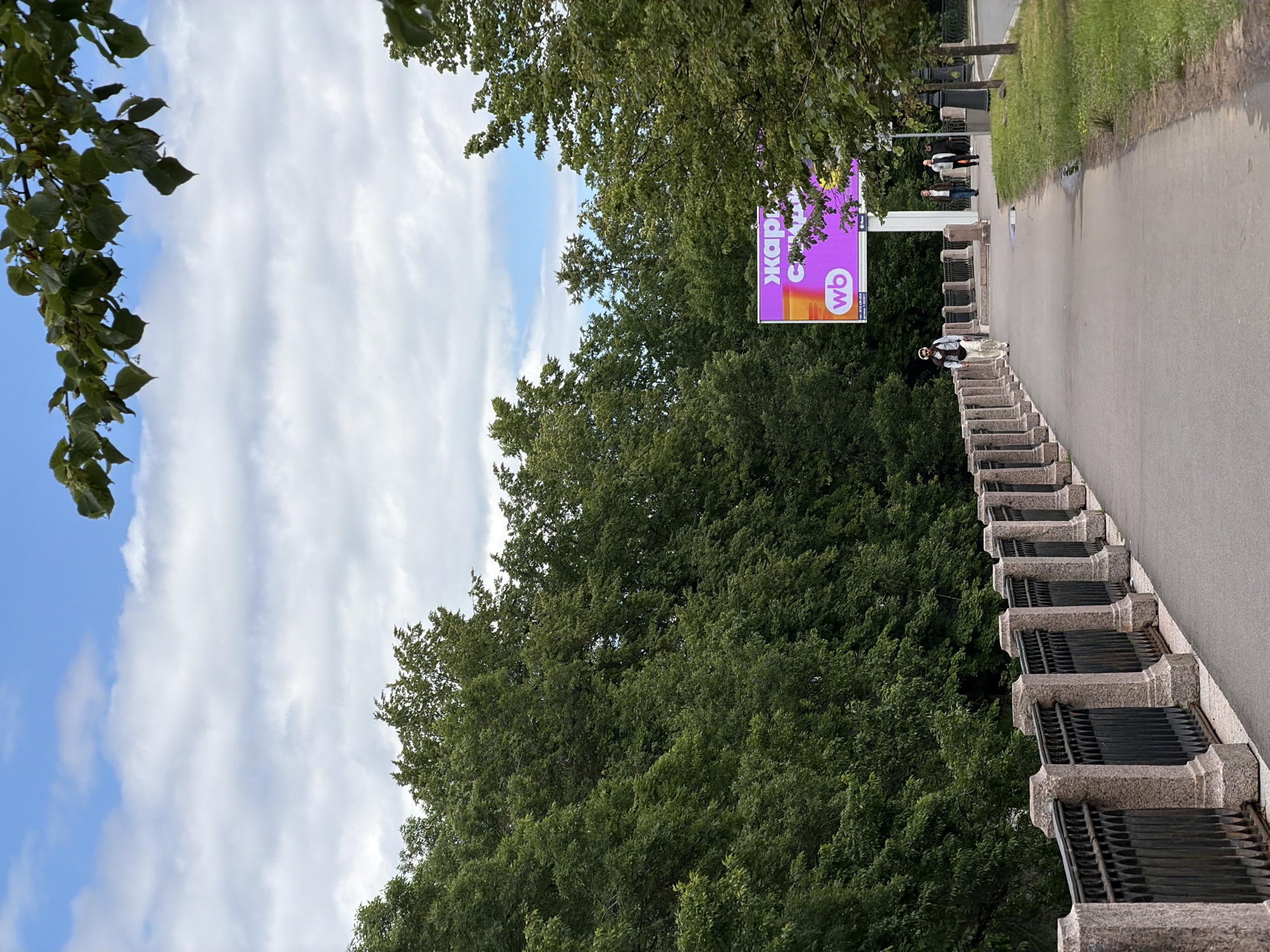The image depicts an outdoor scene with lush green trees dominating the frame. A bright purple billboard with white and orange text is visible in the background, featuring the letters "wb" in a white circle and Cyrillic text that appears to read "жаркий" or "жаркие". A paved walkway runs along the right side of the image, bordered by a grassy area. In the foreground, a person in casual attire is seen walking up a set of granite stairs with metal grates. Further down the walkway, a small group of people are walking away from the camera. The sky is a mix of blue with scattered white clouds, suggesting a partly cloudy day. The overall atmosphere appears relaxed and casual, possibly a park or a public outdoor space in Saint Petersburg, Russia. The lighting suggests it's daytime.