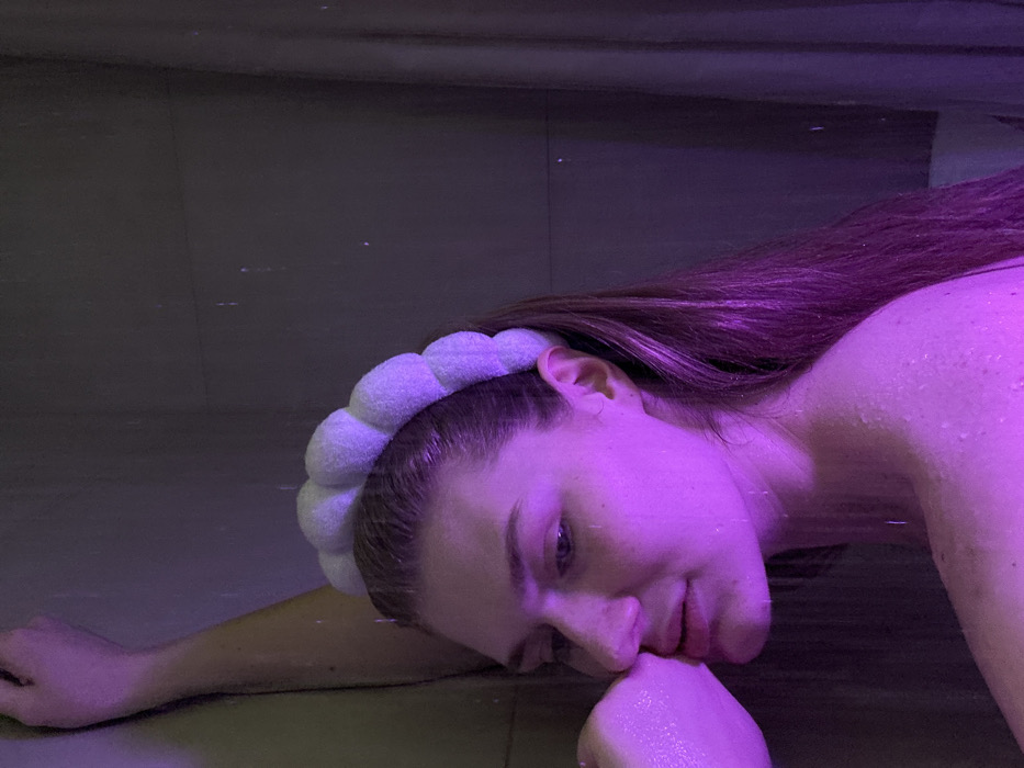 A fair-skinned woman with long, brown hair and a puffy white headband leans against a surface, possibly a pool edge, in a purple-toned light. Water droplets glisten on her skin. She looks towards the camera with a slight smile, resting her cheek on her hand. The scene is dominated by the cool, violet illumination, giving a dreamlike quality.