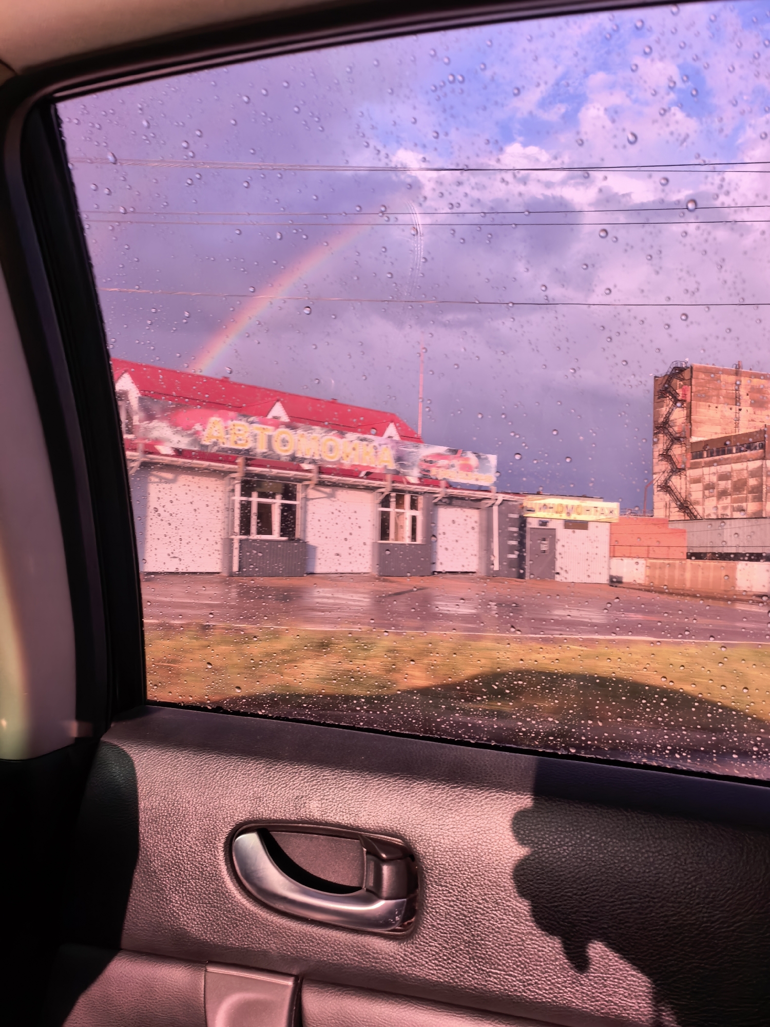 This image captures a view through a car window in Sterlitamak, Russia, on a rainy day. The window is covered in water droplets, creating a blurred effect on the scene outside. Through the window, a rainbow is visible in the purple-hued sky, arcing over a building complex. This complex features a prominent red-roofed structure with the word "АВТОМОЙКА" (car wash) in yellow lettering. Adjacent to it are other buildings, including one with a visible metal staircase. The foreground of the image shows the interior of the car, including a door handle and the textured surface of the car door panel. A shadow, possibly of a person's hand, is cast on the door panel. The overall impression is one of a slightly overcast and wet day, with a touch of color provided by the rainbow.