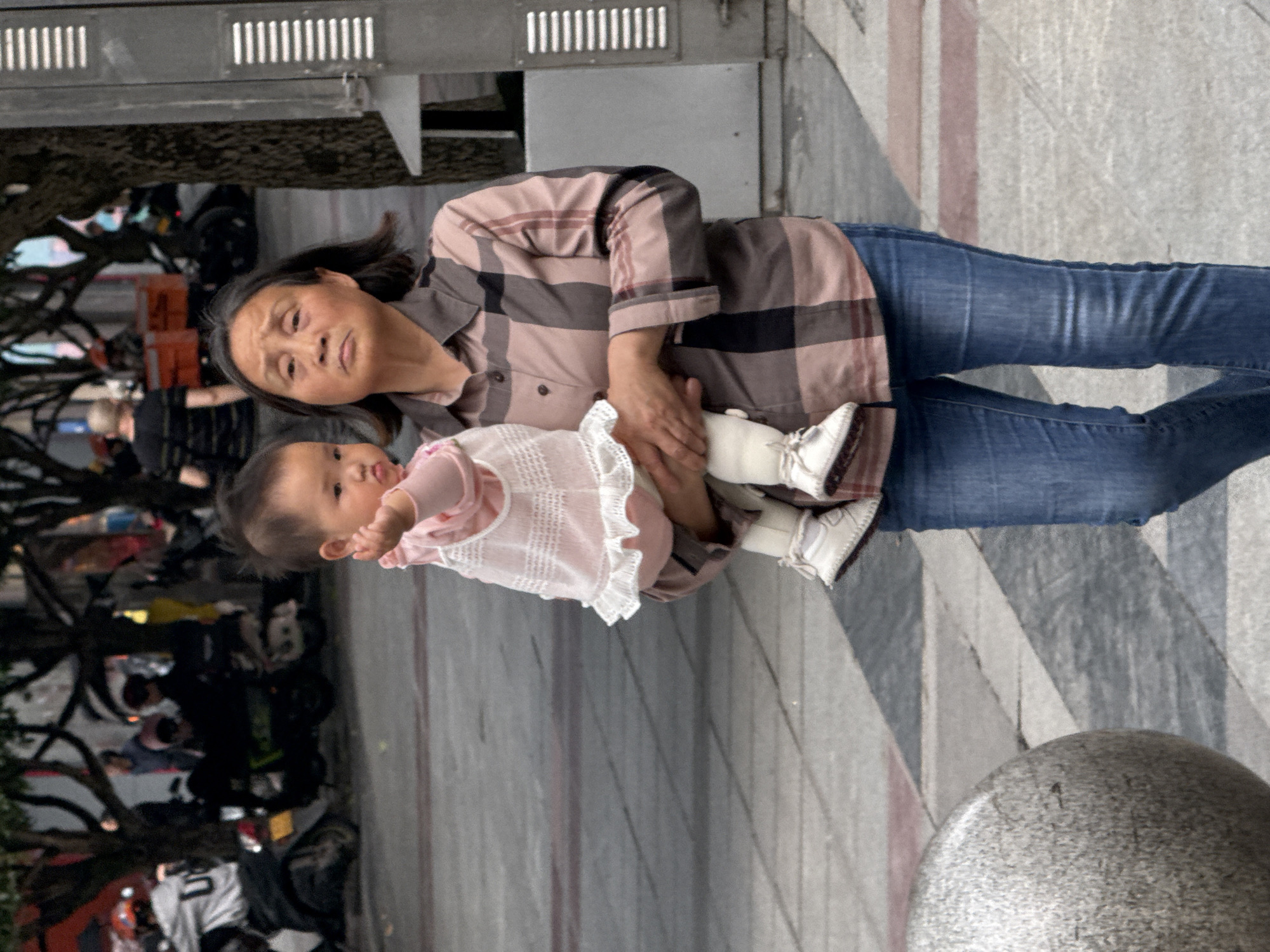 An elderly Asian woman holds a baby on a city street. The woman has short black hair, fair skin, and is wearing a plaid shirt and blue jeans. She looks up and to the left. The baby is wearing a pink shirt and white booties. They appear to be near a street in Jiangjin, China, as evidenced by the Chinese architecture, trees, people, and scooters in the background. The street is paved with light gray concrete or tile and has light pink stripes. A stone or concrete bollard is visible in the lower right corner. There are blurred figures and objects in the background suggesting activity and movement.
