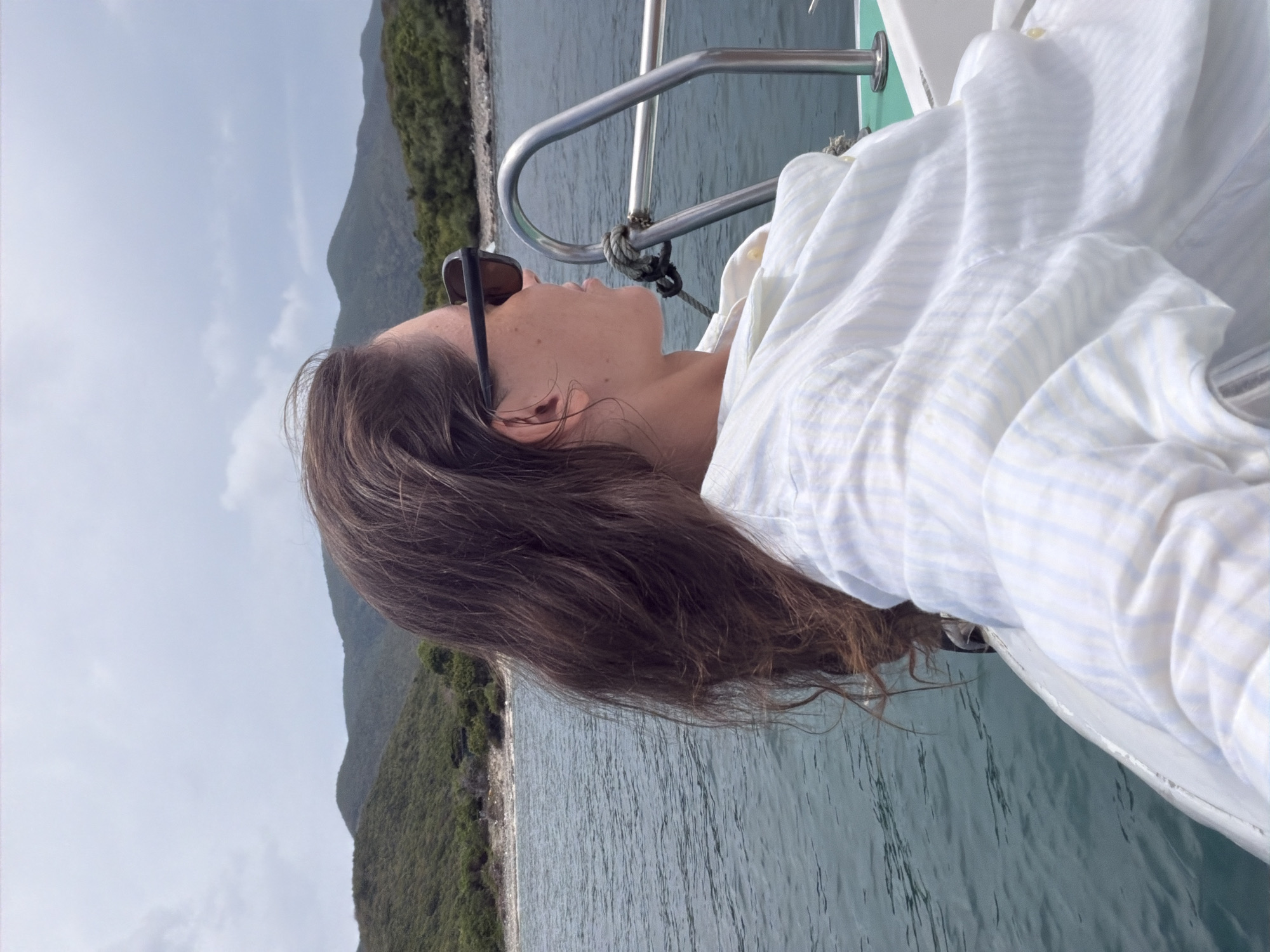 A woman with brown hair and sunglasses reclines on the bow of a boat. She is wearing a light blue and white striped shirt. The boat is traveling on the water near a densely vegetated shoreline. The location is Bac Nha Trang Ward, Vietnam. The railing of the boat is visible. The water is calm. The sky is overcast.