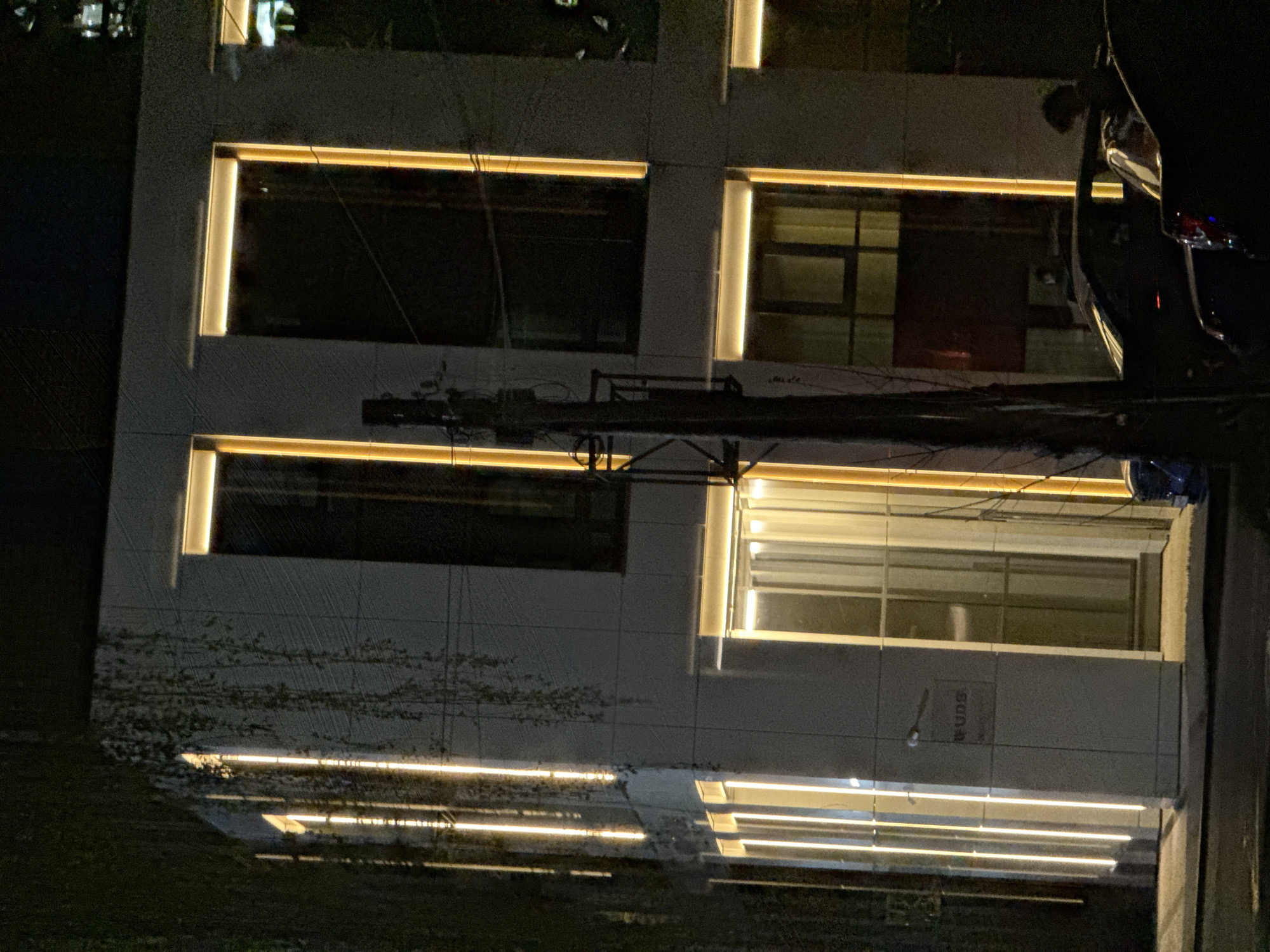 At night, an exterior shot shows a building with rectangular windows, illuminated by lights within the window frames. The building's facade is gray. In the foreground, utility wires crisscross the scene, and a portion of a dark-colored car is visible on the right. The street level is dark, suggesting it is nighttime. The location appears to be Izhevsk, Russia, inferred from the visible Cyrillic lettering on the building's facade. No people are visible.