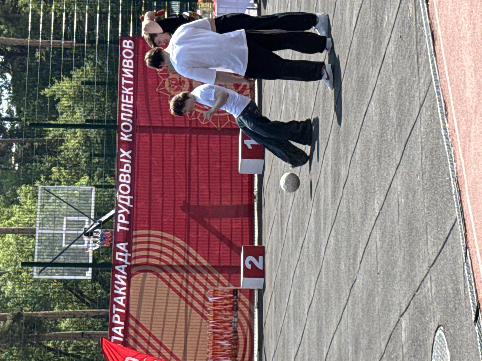 Here is a detailed description of the image:
**Location:** The image appears to be taken at an outdoor sports complex in Dmitrov, Russia. This is suggested by the writing on the banner and the general architecture.
**Subjects:** There are three people visible in the image. Two appear to be leaning back, while the third appears to be kicking a soccer ball that is on the ground in front of them.
**Objects:**
* A soccer ball.
* A basketball hoop, partially obscured by trees.
* A banner with Russian writing, likely advertising a sports event or competition.
* Numbered podiums (1,2,3) are placed on a grey paved surface.
* Trees and a forest-like environment in the background.
**Action:** The third person is kicking a soccer ball. The other two appear to be observing, relaxing, or potentially part of a team activity.
**Environment:** The scene is outdoors, likely on a sports field or paved area designed for athletic activities. The lighting suggests it is a bright, possibly sunny, day. The background includes trees, indicating the location is near a park or wooded area.