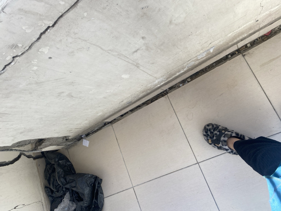 The image displays a damaged interior corner in Khan Yunis, Palestinian Territory, featuring a light-tiled floor and two concrete walls. Both walls exhibit significant structural compromise, with deep, jagged cracks evident at their intersection and extending across their surfaces. A prominent vertical and horizontal separation is visible at the corner, exposing underlying construction materials and debris. A narrow, debris-filled gap runs along the base of the right wall. A crumpled black plastic bag is situated in the far-left corner, adjacent to small pieces of white paper. In the lower frame, a person's left foot is visible on the tiled floor, wearing a black and white leopard-print fur slipper with a dark strap and gold-colored buckle, paired with dark trousers and a visible portion of a light blue garment.