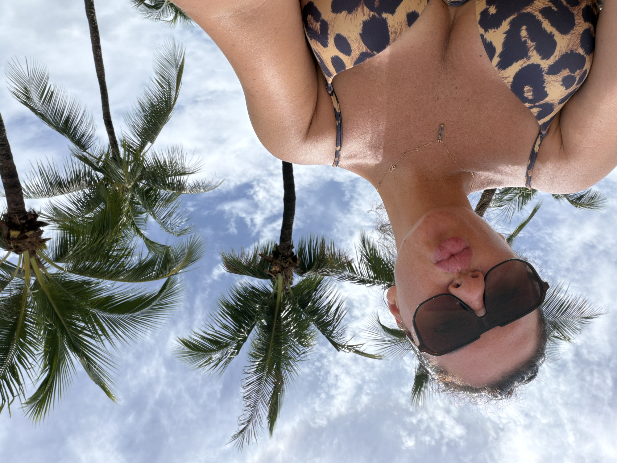 A woman wearing sunglasses and a leopard-print bikini top poses for a selfie outdoors. She has her lips puckered, and is wearing a gold necklace. Palm trees and a cloudy blue sky are visible in the background. The location is Bang Muang, Thailand.