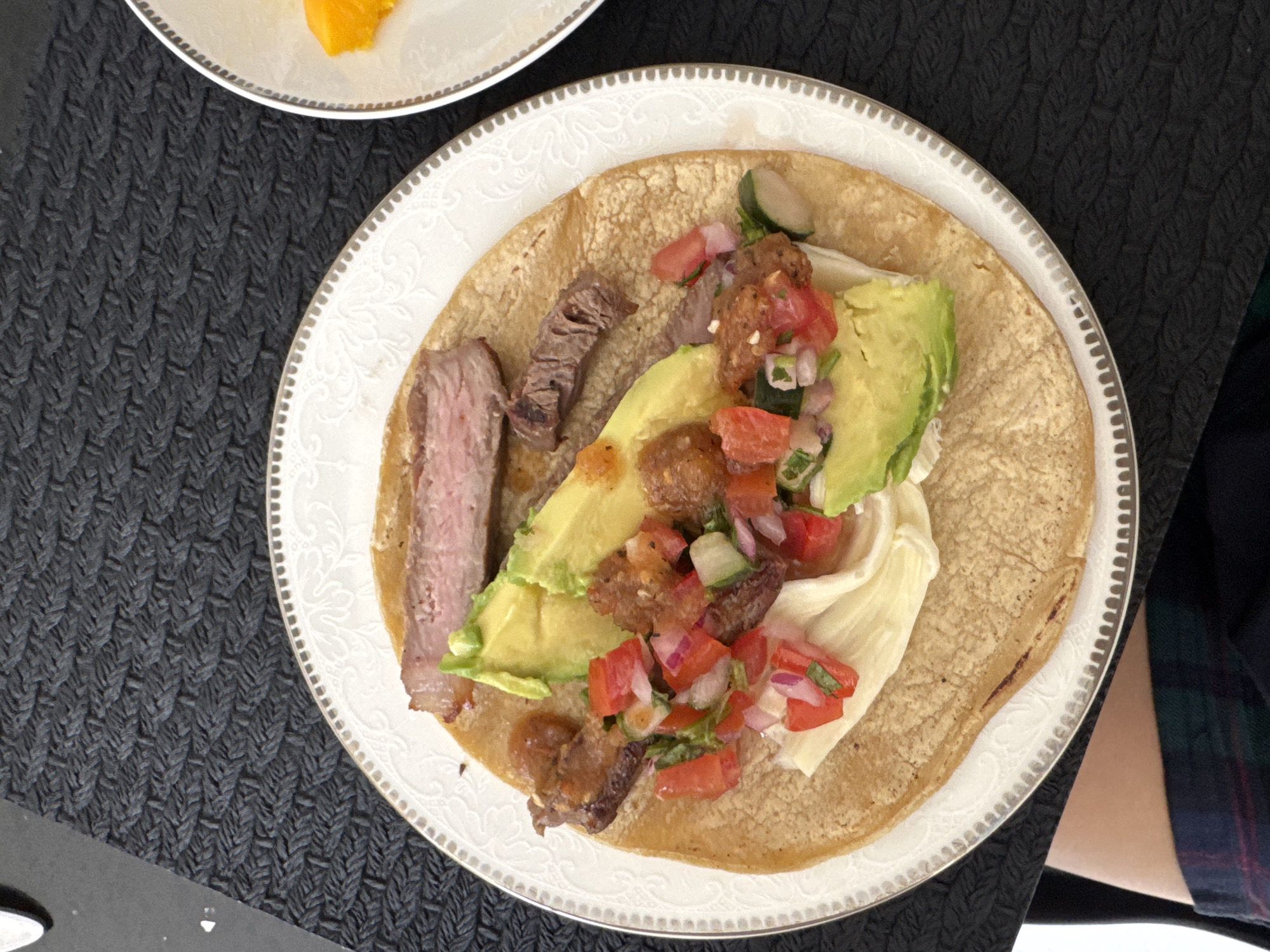 A top-down shot reveals a well-adorned plate holding a single taco, its ingredients arranged attractively. Slices of what appears to be steak flank a bed of creamy avocado, complemented by a vibrant salsa of tomato, red onion, and herbs, all atop a soft tortilla. The plate itself boasts a decorative floral pattern around the rim. To the side, the corner of another plate bears a segment of orange-colored food. The scene sits on a textured black surface, possibly a placemat, while a glimpse of a plaid-clad arm suggests someone is seated nearby. The location is not discernible from the image.