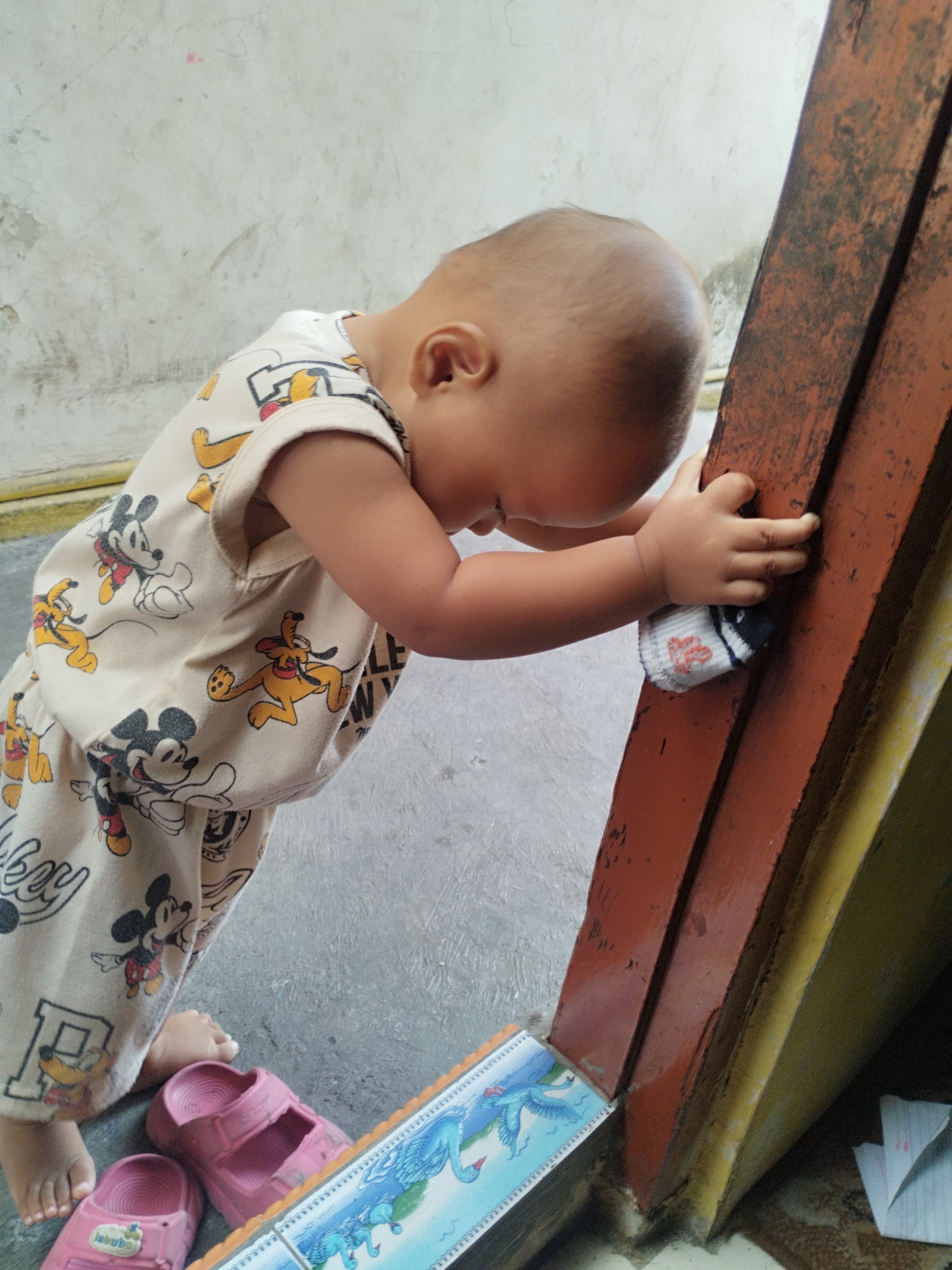 A toddler, wearing a light-colored onesie with a cartoon character print, leans against a weathered, reddish-brown wooden post, holding a small, white sock with red accents in their right hand. The toddler's head is bowed, and their face is not visible. The background consists of a rough, off-white wall and a grey concrete floor. Pink sandals are visible on the floor near the toddler's feet. To the right of the wooden post, a yellow structure is partially visible. A colorful printed surface, possibly a piece of paper or cardboard with a depiction of mythical creatures, is also on the floor. The scene appears to be an outdoor area, possibly a residential yard or alleyway in Surabaya, Indonesia, given the context. The lighting suggests daylight.