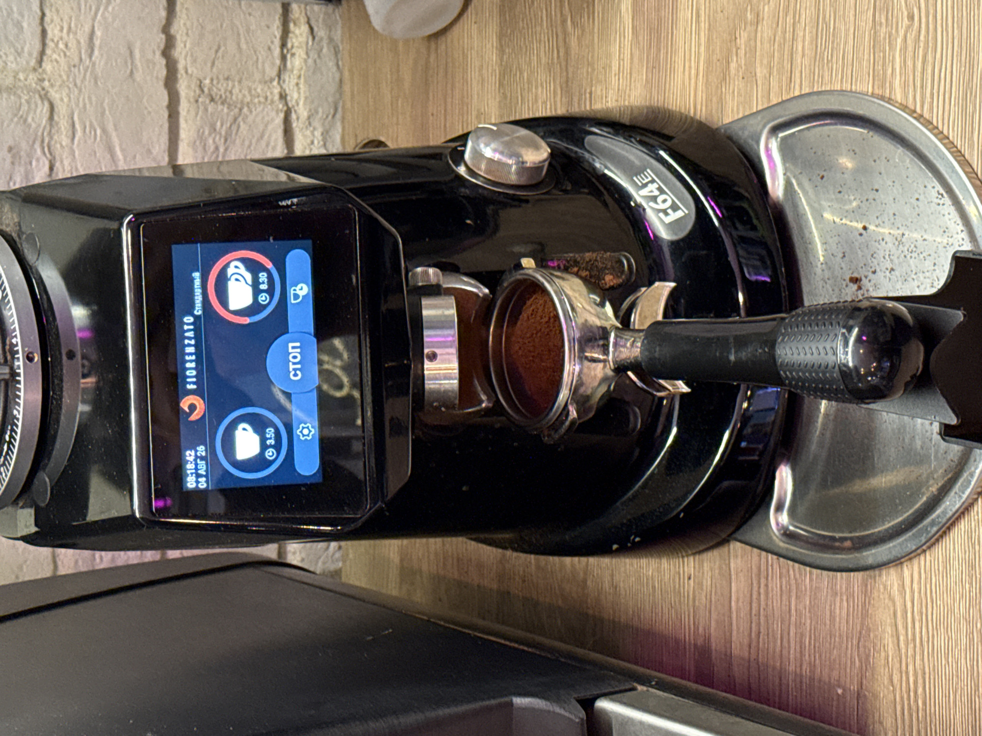 The image shows a close-up of a black Fiorenzato F64E coffee grinder, likely in a cafe setting. The grinder has a digital display showing the time as 08:18:42 and the date as 04 AUG 20. It also displays two coffee cup icons with associated values, one showing "3.50" and the other "6.30", suggesting programmable doses. A portafilter filled with ground coffee is inserted into the grinder mechanism, and a stainless steel drip tray is visible below. The background includes a brick wall and a wooden surface. The grinder is positioned on a wooden counter. There is no direct evidence of the specific location in Moscow, Russia, other than it is an indoor commercial setting.