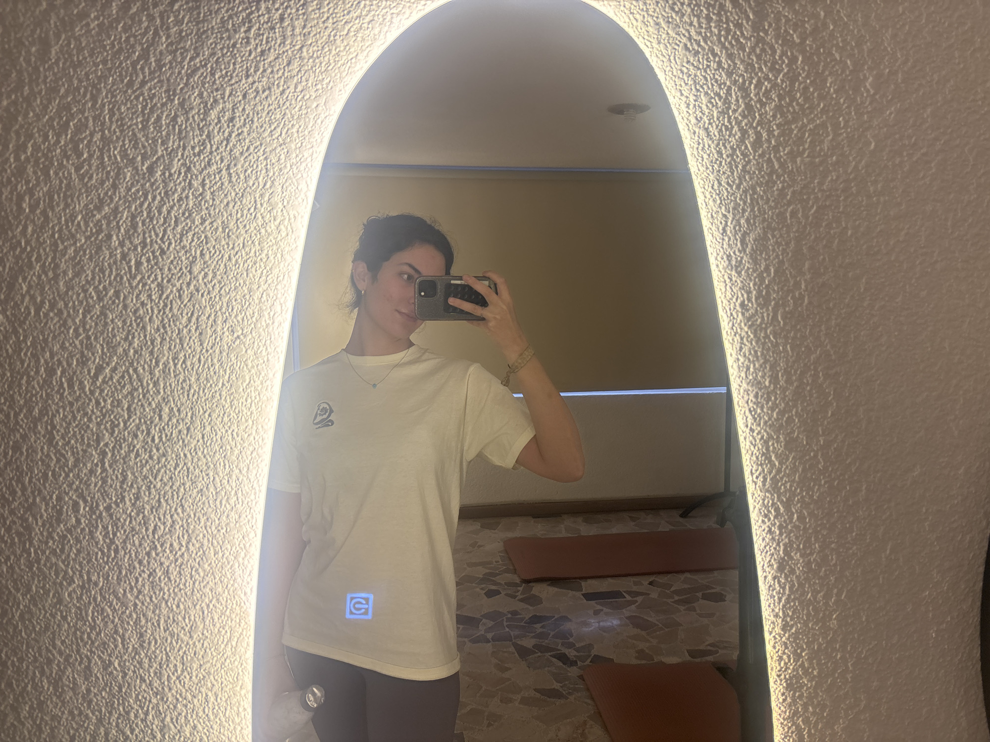 A young woman stands in front of a well-lit arched mirror, likely taking a selfie. She has dark hair pulled back, a light-colored t-shirt, and grey leggings. She holds a phone up to her face.
The reflection reveals a room with tiled floors and yoga mats laid out, suggesting a yoga studio. The background shows a window with a shade partially drawn. The location may be in Puebla, Mexico, based on available information.