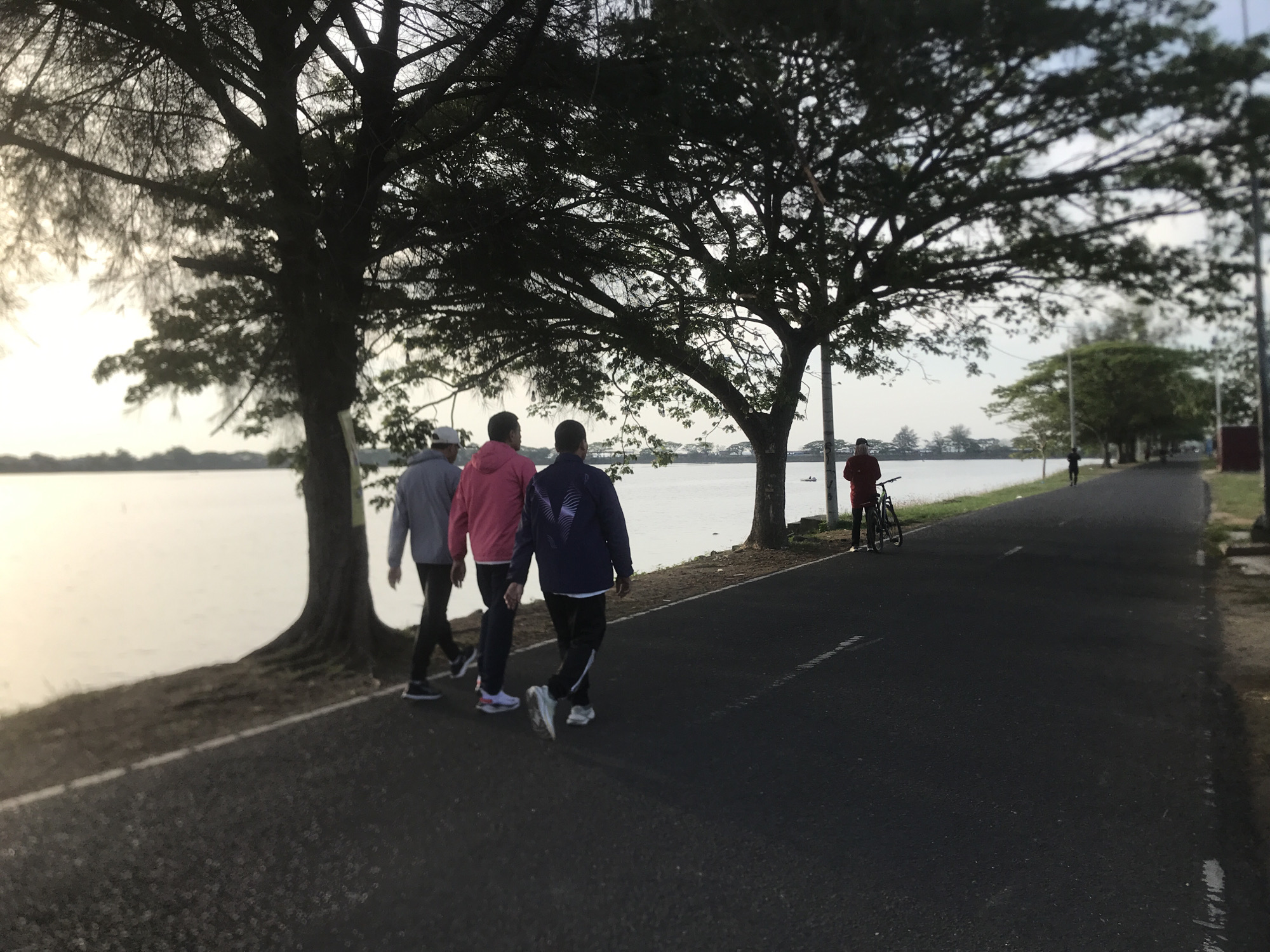 The image captures an outdoor scene at what appears to be a lakeside or riverside path in Lhokseumawe, Indonesia, likely during early morning or late afternoon given the soft, bright light and hazy sky.
In the foreground, three men are walking away from the camera on a paved path that runs parallel to a large body of water. The man on the left wears a grey hooded top, the middle man a pink hooded top, and the man on the right a dark blue jacket with a light-colored design on the back. They are dressed in casual activewear, suggesting a leisurely walk or exercise.
Large, dark-foliaged trees line the left side of the path, their branches stretching over the water and creating a natural canopy. These trees are silhouetted against the bright, reflective surface of the water and the hazy sky.
Further down the path on the right, another person in a red top stands beside a bicycle. In the far distance along the path, a faint silhouette of another individual can be seen walking. The path itself is dark asphalt with faint white markings.
The setting is serene, with the tranquil water stretching into the distance and faint land or structures visible on the far horizon. The overall impression is one of peaceful outdoor activity in a scenic natural environment. No visible text is present in the image.