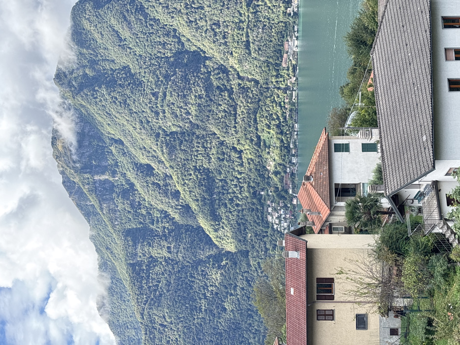 This image presents a panoramic view of the landscape in Nesso, Italy, specifically showcasing a section of Lake Como and its mountainous surroundings.
In the foreground, an arrangement of residential buildings descends a slope. The most prominent structures include a two-story beige building with a terracotta tiled roof, brown window frames, and partially visible dark brown shutters. To its right, another white building with a terracotta tiled roof features green shutters on its windows and a balcony opening. Further down and partially obscured, a third, smaller white building with a dark grey tiled roof is visible, featuring an external concrete staircase with railings leading to an upper landing. These buildings are interspersed with dense green vegetation, including trees and shrubs. No individuals are present or visible in this section.
The midground is dominated by a wide expanse of teal-colored water, identified as Lake Como, exhibiting subtle ripples across its surface. Along the distant shoreline, numerous light-colored residential and commercial structures are clustered at the base of the mountains.
The background is characterized by a series of steep, imposing mountains heavily covered in dense green foliage, indicating extensive tree growth. The upper slopes and peaks of these mountains are partially obscured by white and light grey clouds. The sky above is partly cloudy, with patches of blue sky visible between the white and grey cloud formations. The scene is illuminated by natural daylight.