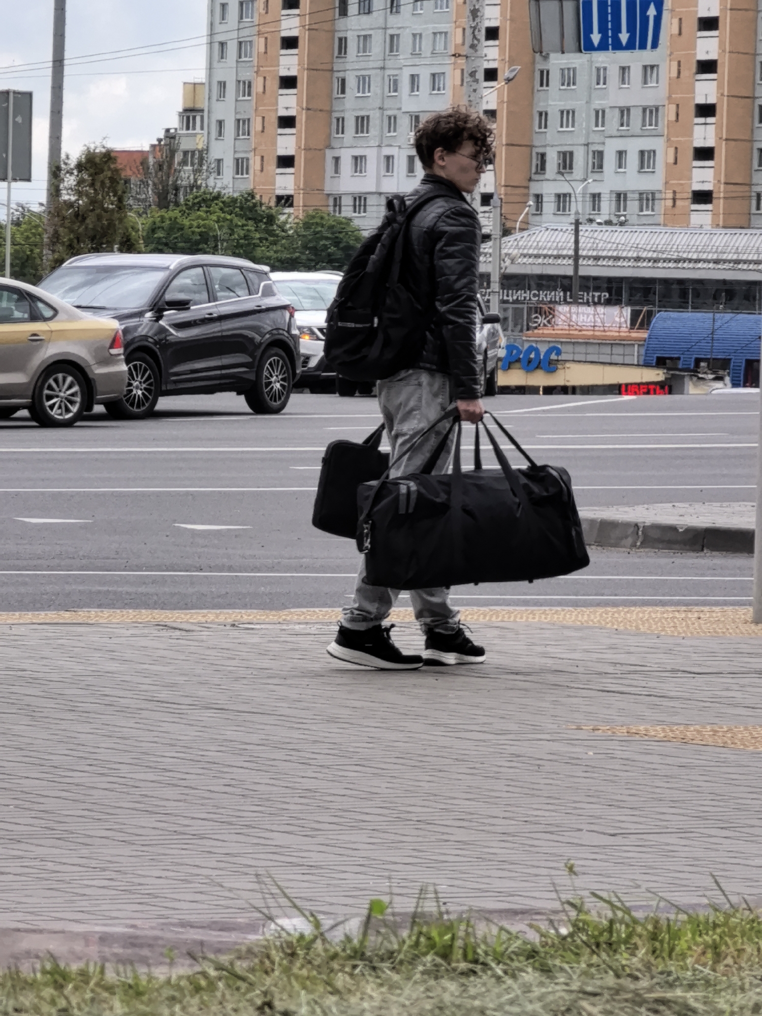 A young man with curly brown hair and glasses, wearing a black leather jacket, grey jeans, and black sneakers, stands on a sidewalk. He is carrying two large black duffel bags in his right hand, and a backpack is visible on his left shoulder. The man is looking to his right, away from the camera.
In the background, there is a street with several cars, including a dark grey SUV and a yellow sedan. The cars are driving or stopped on the road. Behind the street, there is a large, multi-story building with a grey and beige facade and numerous windows. The sky appears overcast.
A sign with Cyrillic text is visible on the building, reading "ЦИЯНСКИЙ ЦЕНТР" and below it, "POC". Another sign below that says "ЦВЕТЫ" in red letters. These signs suggest a commercial area, possibly a shopping center or a service establishment. The overall scene appears to be an urban environment during the daytime, under cloudy weather conditions.