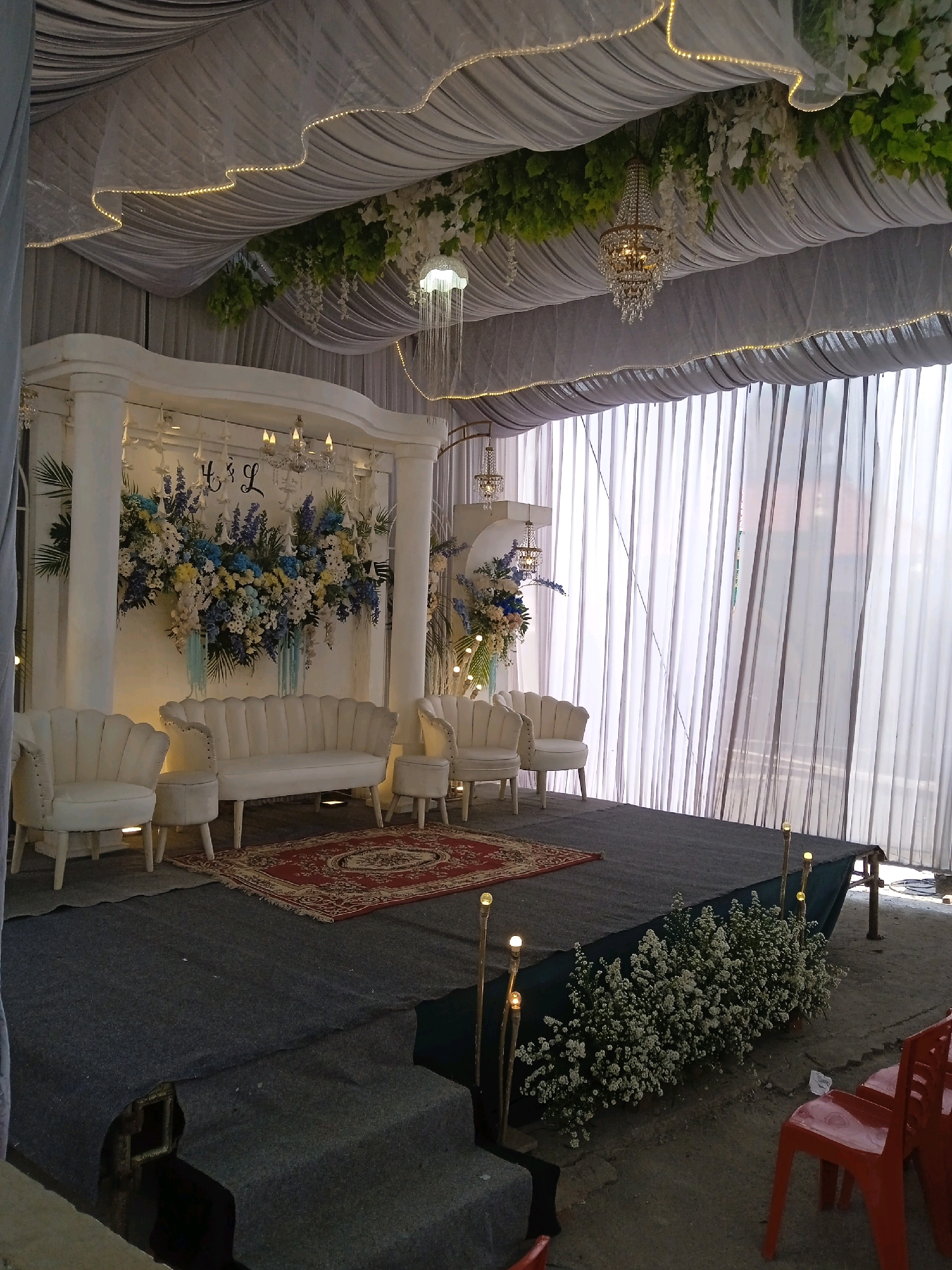 The image depicts a decorated outdoor venue in Kelurahan, Indonesia, likely set up for an event such as a wedding or reception. A draped white tent ceiling with integrated string lights and hanging greenery creates an ambiance. A central stage area features a white backdrop with floral arrangements and the initials "H & L" on display. White upholstered furniture, including a sofa and armchairs, are arranged on the stage. A patterned rug is placed in front of the furniture. The stage is carpeted in grey, with steps leading down. To the right of the stage, decorative lights on sticks illuminate the foreground. The background consists of sheer white curtains. Red plastic chairs are visible in the lower right corner of the frame. The ground outside the stage area appears to be rough terrain. No people are visible in the image.