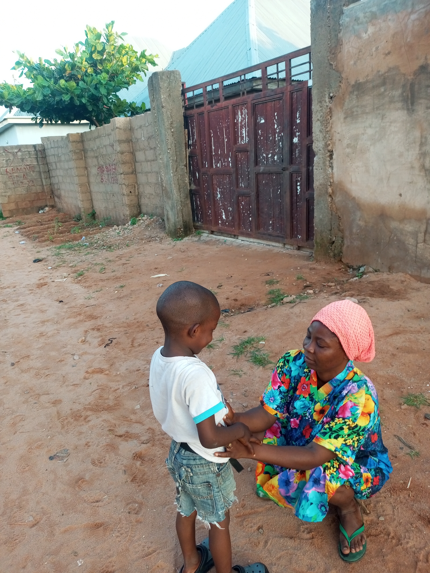 In the city of Tempure, Nigeria, a young boy wearing a white t-shirt and distressed denim shorts stands on a dirt path, facing a woman. The woman, wearing a pink headscarf and a vibrantly colored floral dress, is crouched down and holding the boy's hands. Behind them, a rusty metal gate stands in front of a textured wall. To the left, a brick wall with graffiti is visible, topped with green foliage. The ground is covered in reddish-brown dirt, with scattered debris and footprints.