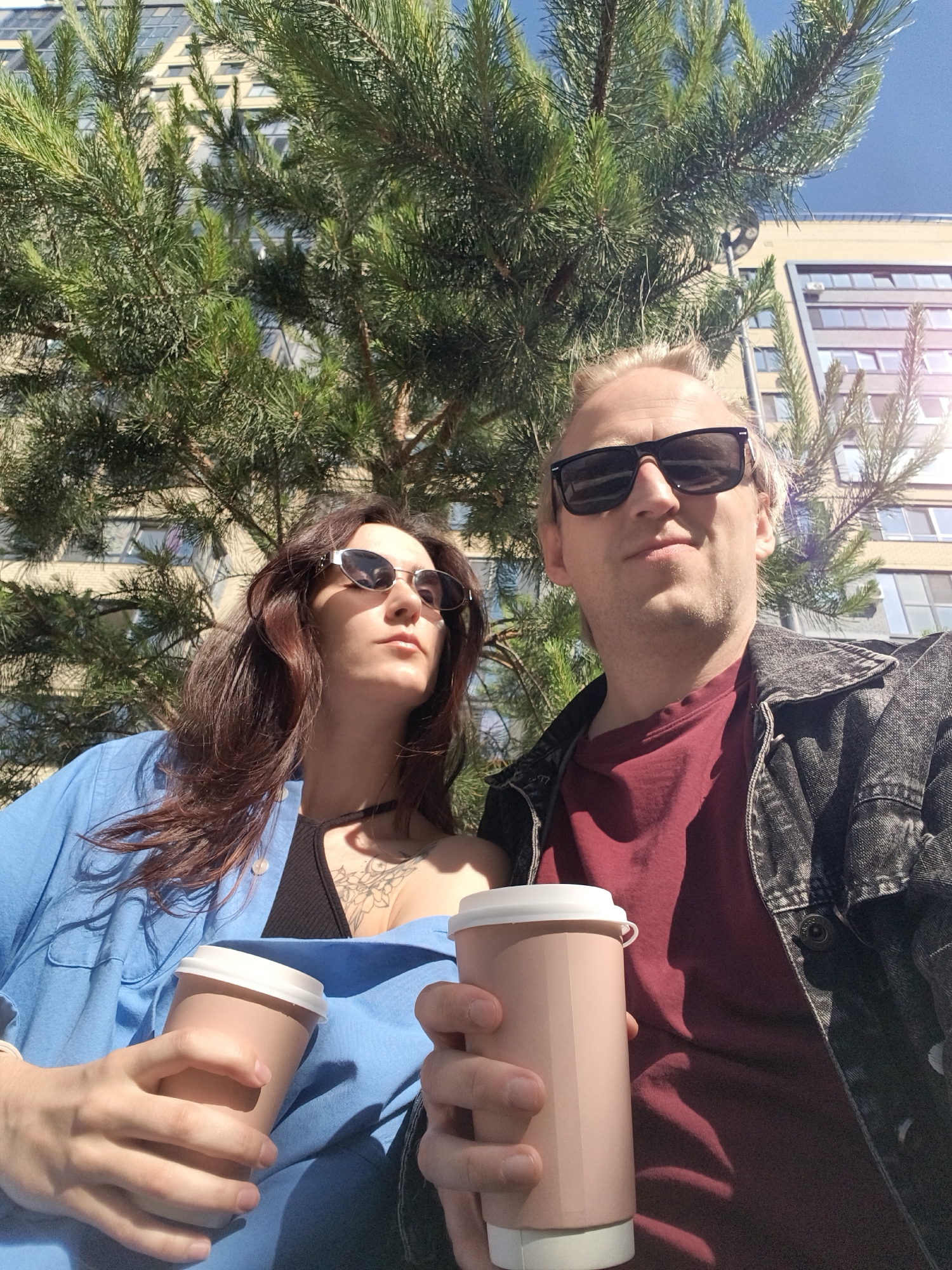 A man and a woman are captured in a bright, high-angle selfie outdoors in Tyumen, Russia. The man, on the right, has blonde hair and wears dark rectangular sunglasses, a dark red t-shirt, and a dark grey denim jacket. He looks directly at the camera with a relaxed expression, holding a light brown disposable coffee cup with a white lid. The woman, on the left, has long brown hair and wears small, oval sunglasses, a light blue button-up shirt over a black top, revealing a tattoo on her chest/shoulder. She looks upwards and to her right, holding a matching coffee cup.
The background is dominated by the vibrant green needles and branches of a pine tree, partially obscuring a modern multi-story building with numerous windows. A clear blue sky is visible in the upper right, indicating a sunny day. The strong sunlight creates distinct shadows on the subjects and their cups. The scene conveys a casual outdoor break, likely during the daytime, in an urban or suburban setting.