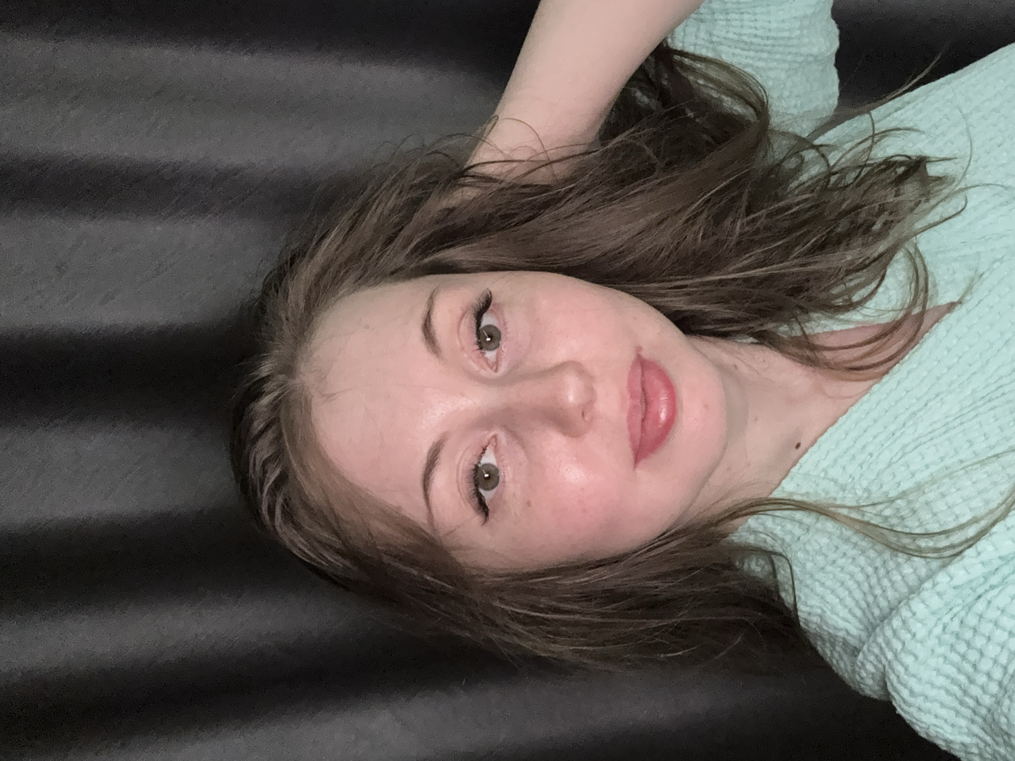 A young woman is depicted in a close-up portrait, lying down with her head tilted to the side, looking directly at the viewer. Her expression is relaxed, with a soft gaze and slightly parted lips, indicating a subtle smile or a state of calm. She has long, wavy, medium-brown hair that is spread around her head, framing her face.
Her skin appears clear, and she wears subtle makeup, including dark eyeliner or mascara highlighting her light brown or hazel eyes, and a glossy pinkish color on her lips. She is wearing a light teal or mint green top with a distinctive waffle-knit texture, visible around her shoulder and the left side of her head. Her right arm is bent and raised, with her hand partially obscured by her hair near the top of her head.
The background is a dark, solid color, featuring subtle horizontal ridges or creases, suggesting a soft, textured surface such as a bed, blanket, or upholstered furniture. The lighting is soft and even, indicating an indoor setting. There is no visible text in the image. The overall impression is one of tranquility and ease.