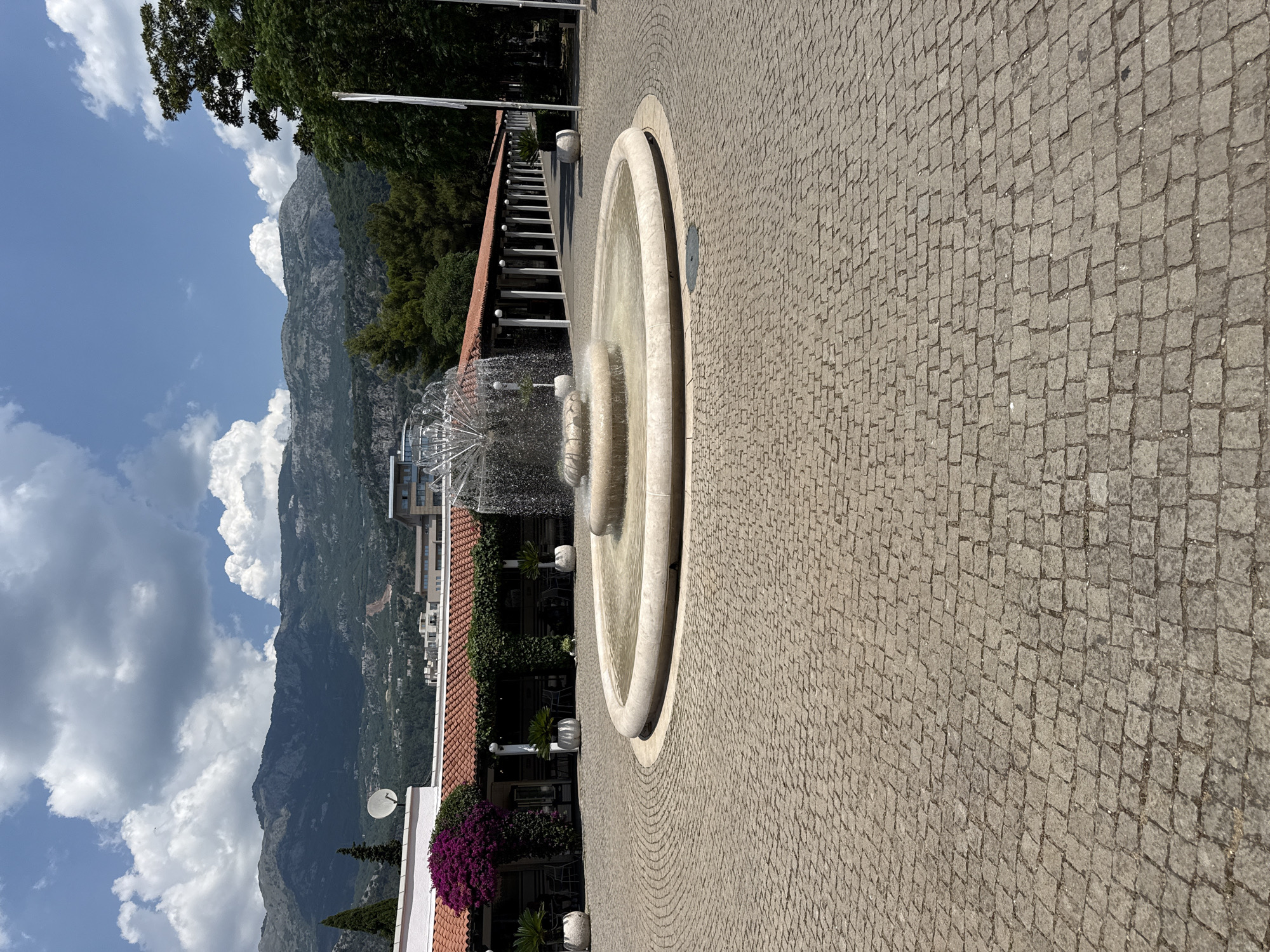 This is a photograph taken in Budva, Montenegro, oriented vertically. The scene depicts a cobblestone promenade leading to a multi-tiered circular fountain with water spraying from its center. Beyond the fountain, a building with a red-tiled roof and white trim is visible, adorned with lush green ivy and a vibrant display of purple flowers. Further back, more modern buildings can be seen. In the distance, majestic mountains rise under a sky filled with fluffy white clouds. The time of day appears to be daytime, with bright sunlight casting shadows. The overall mood is serene and picturesque, suggesting a pleasant day in a scenic coastal town. No people are visible in the frame, and there is no visible text.